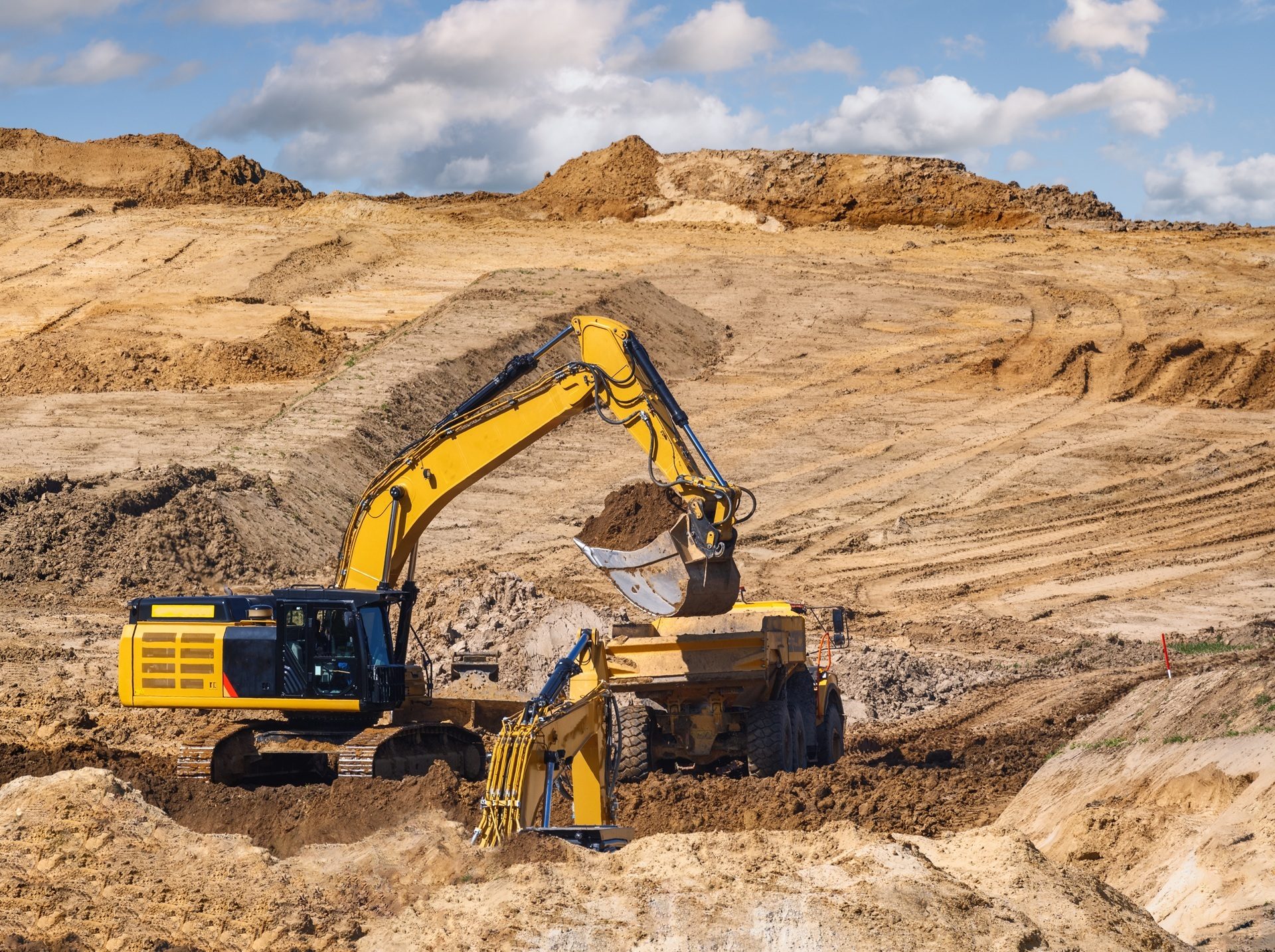 A yellow excavator loads soil into a dump truck at a large construction site under a cloudy sky.