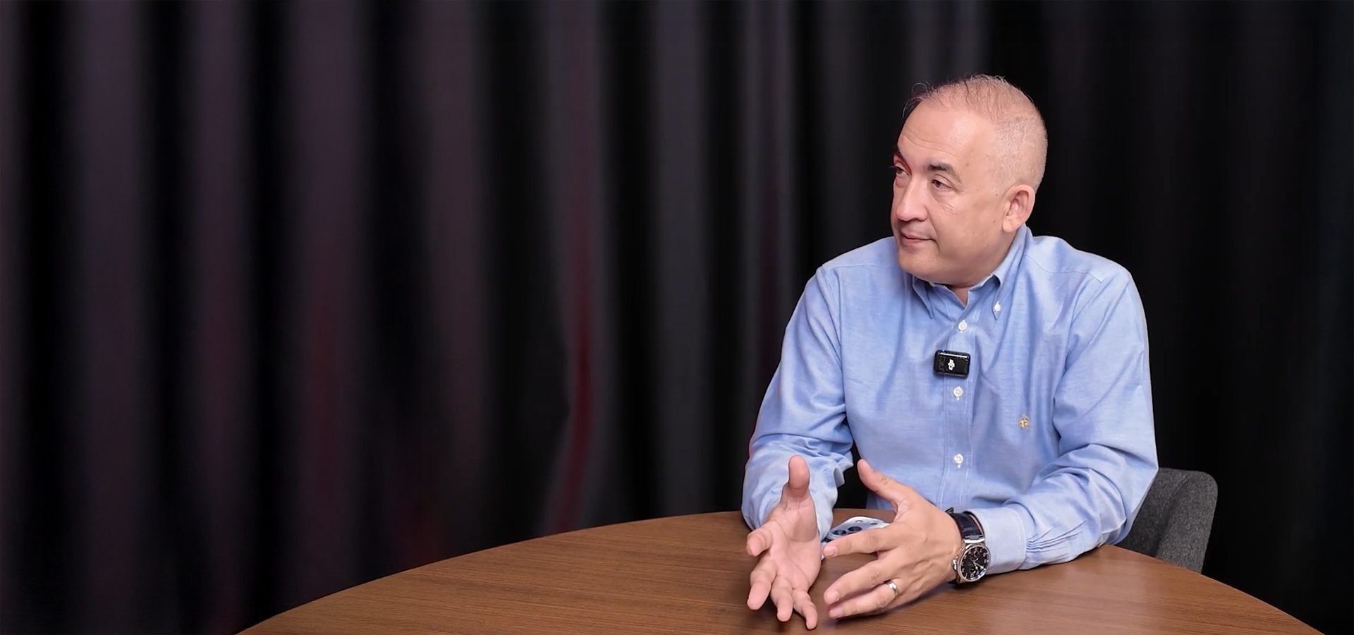 A man in a blue shirt sits at a table, gesturing with his hands, looking left, with a microphone.