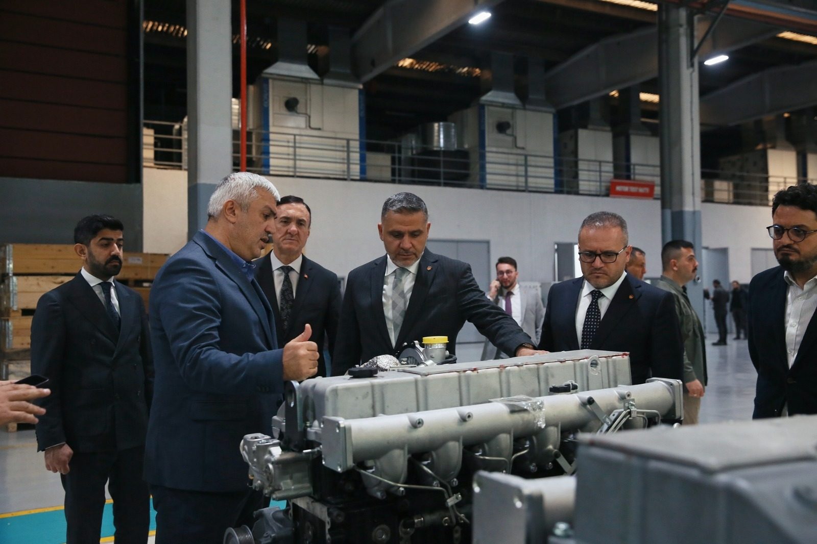 Men in suits inspect a large industrial engine in a factory setting.