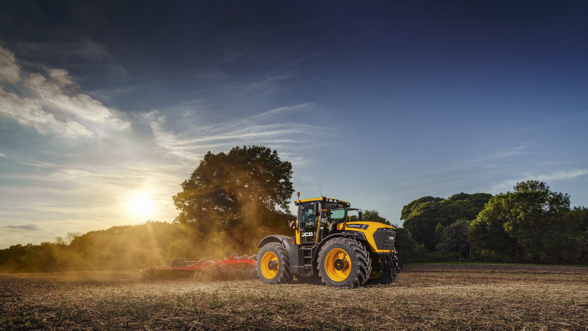 Yellow JCB tractor plowing a field at sunset, with dust rising.