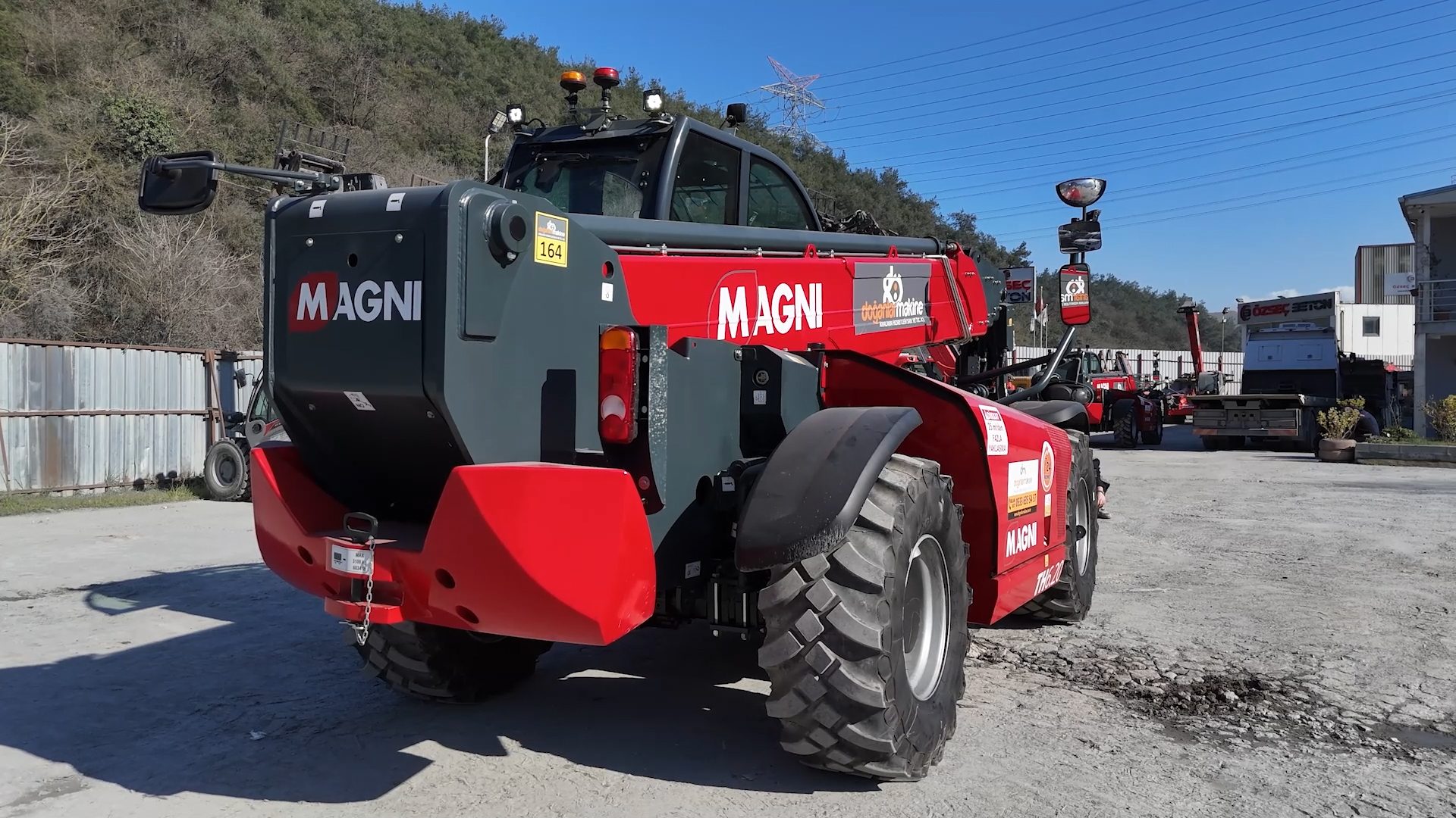 A large red and grey Magni telehandler parked outdoors on a sunny day, viewed from its rear-right.