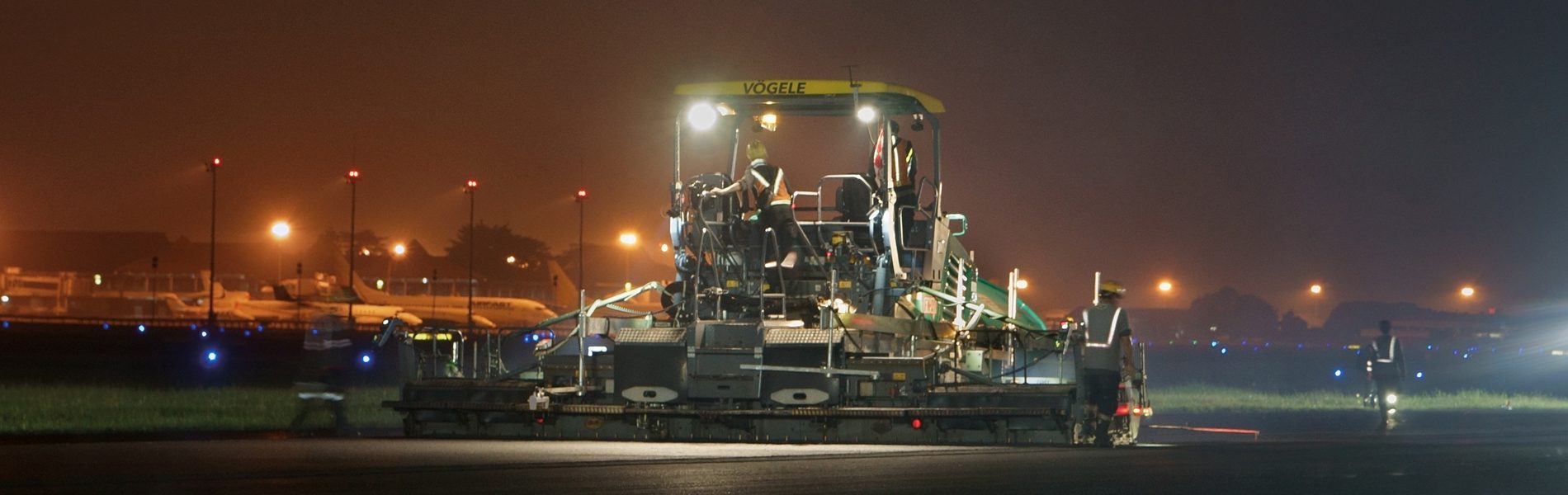 Night airport construction: a large paver machine and workers on a runway, planes in background.
