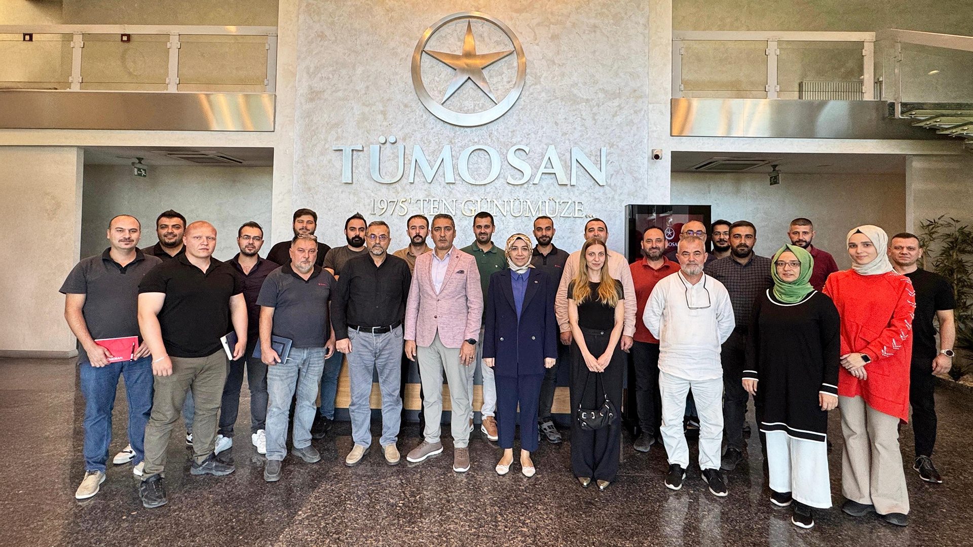 A group of people, mostly men, and some women, pose in front of a TÜMOSAN logo and text.