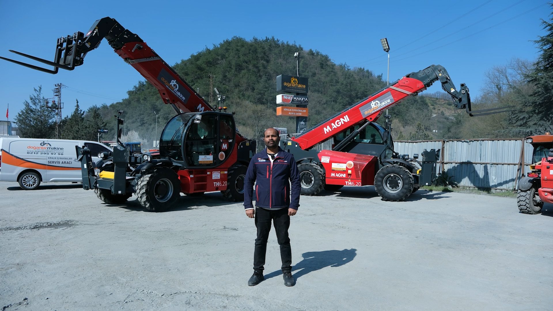 A man stands between two large red Magni TH6.20 telehandlers at a "doğanlar makine" facility under a clear sky.