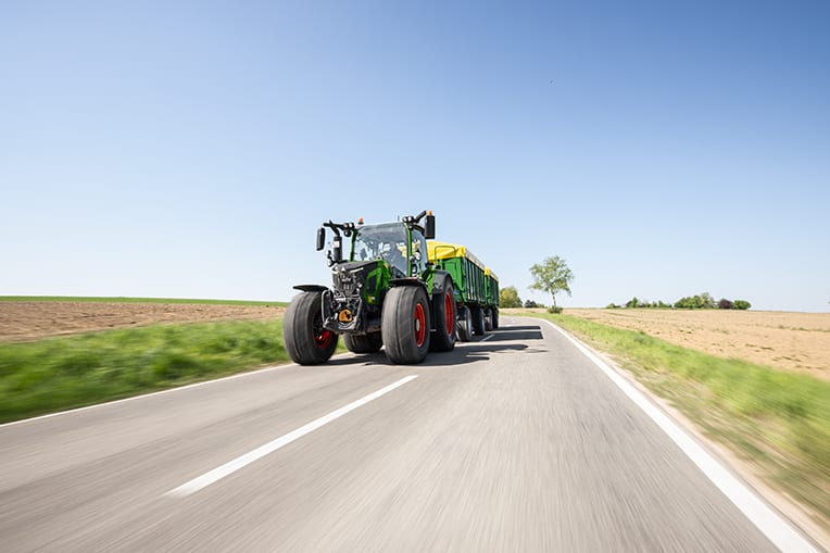 Green tractor pulling trailer on road through fields under clear sky.