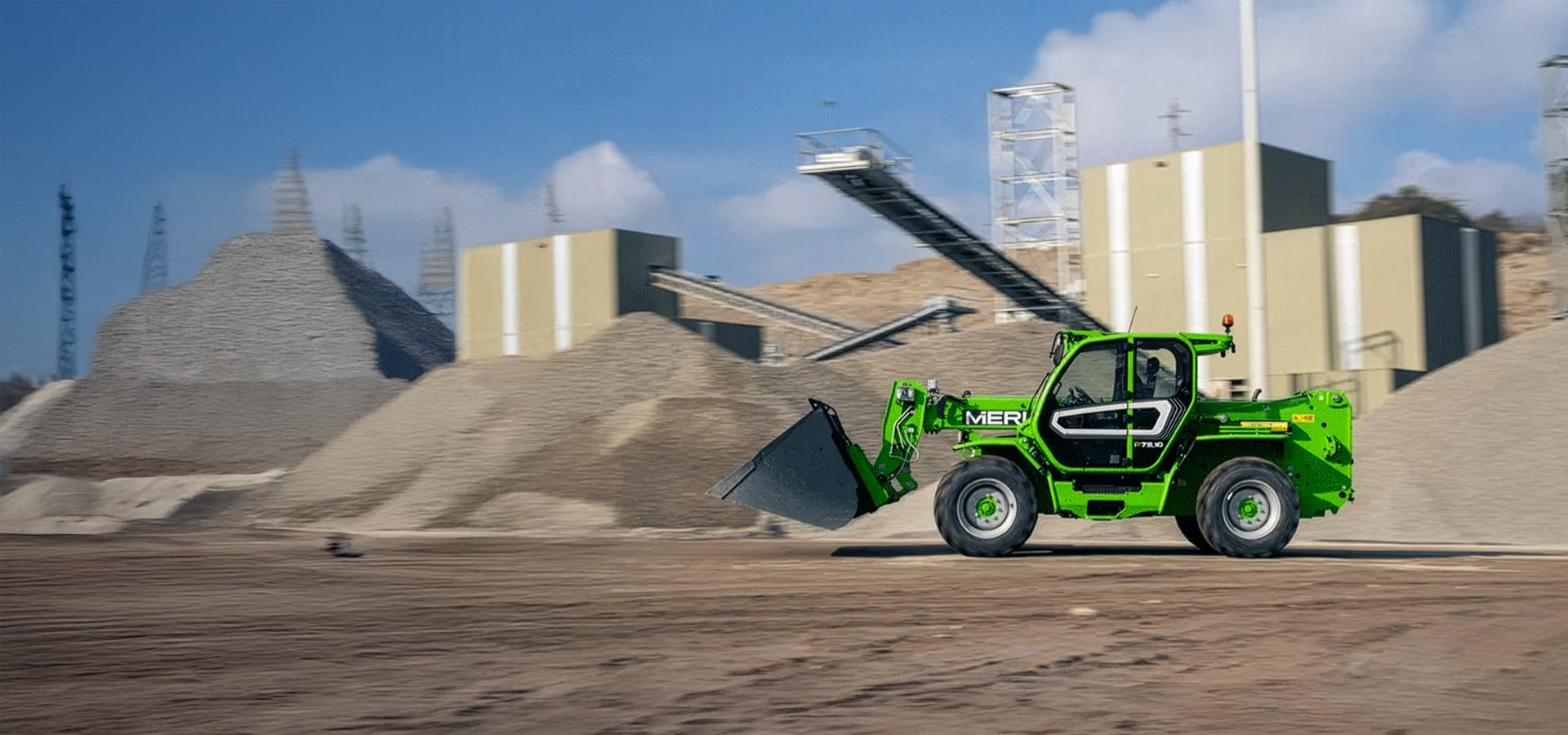 Green Merlo telehandler driving at a quarry with aggregate piles.
