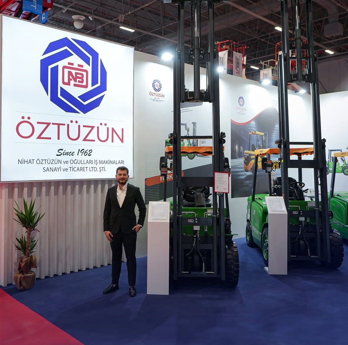 Man at ÖZTÜZÜN's exhibition booth with green forklifts and a "Since 1962" sign.