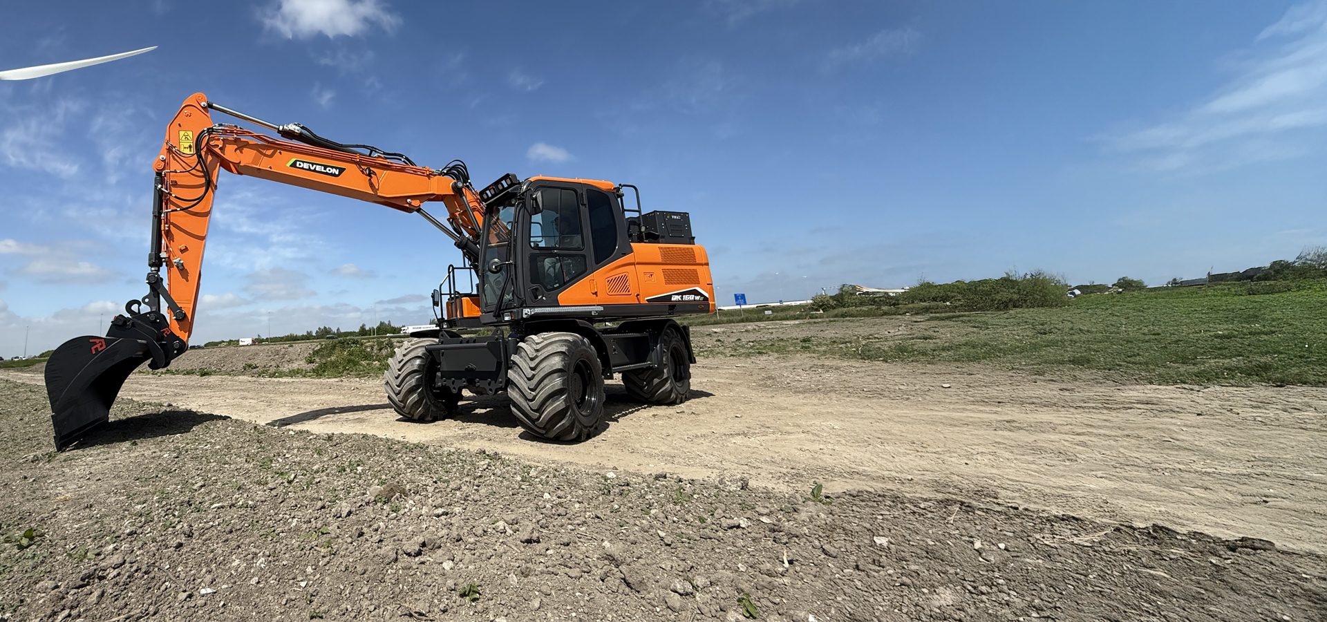 Orange wheeled excavator on dirt under a blue sky, with a wind turbine blade.