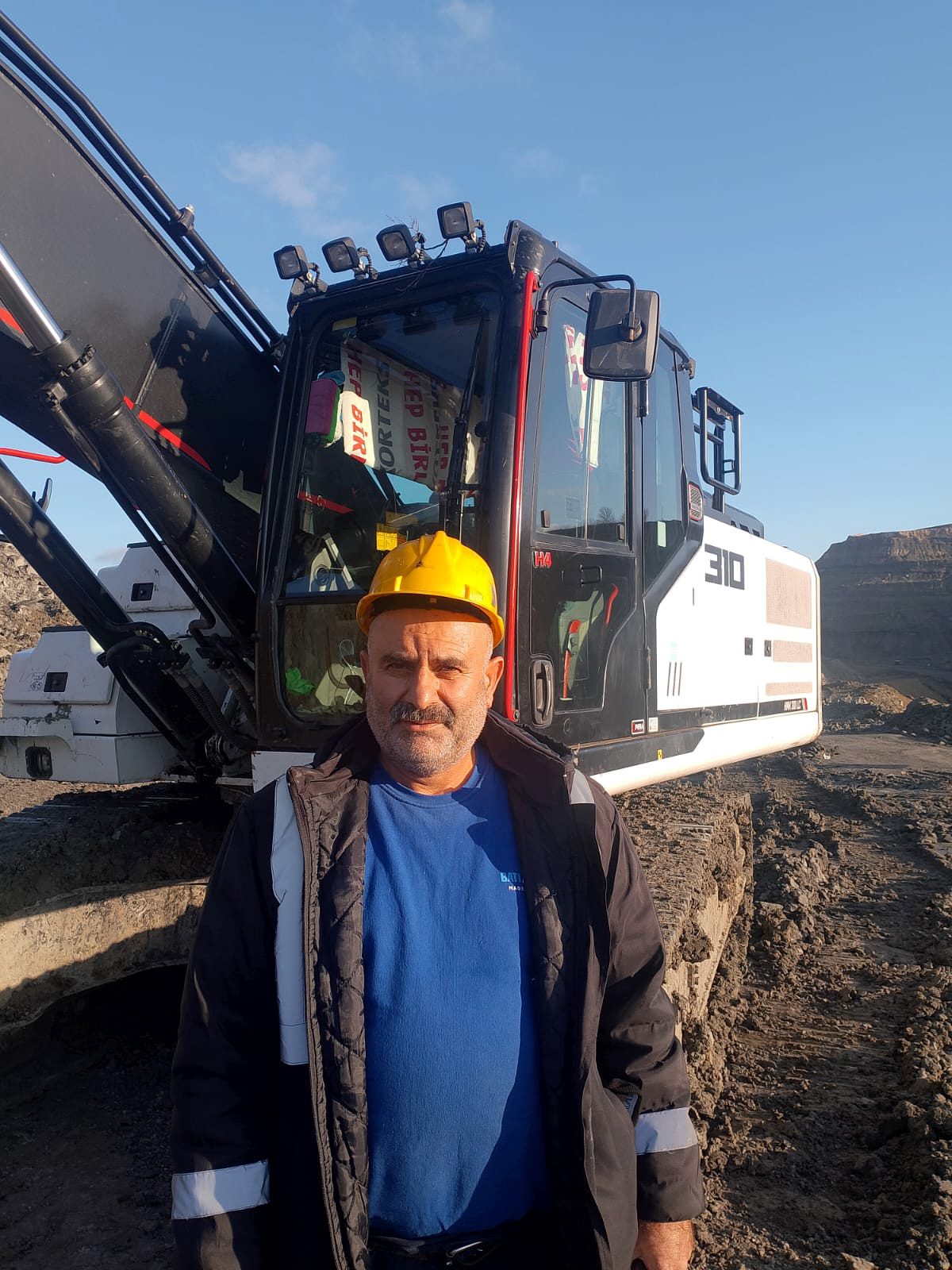 Man in yellow hard hat and blue shirt stands in front of an excavator at a worksite.