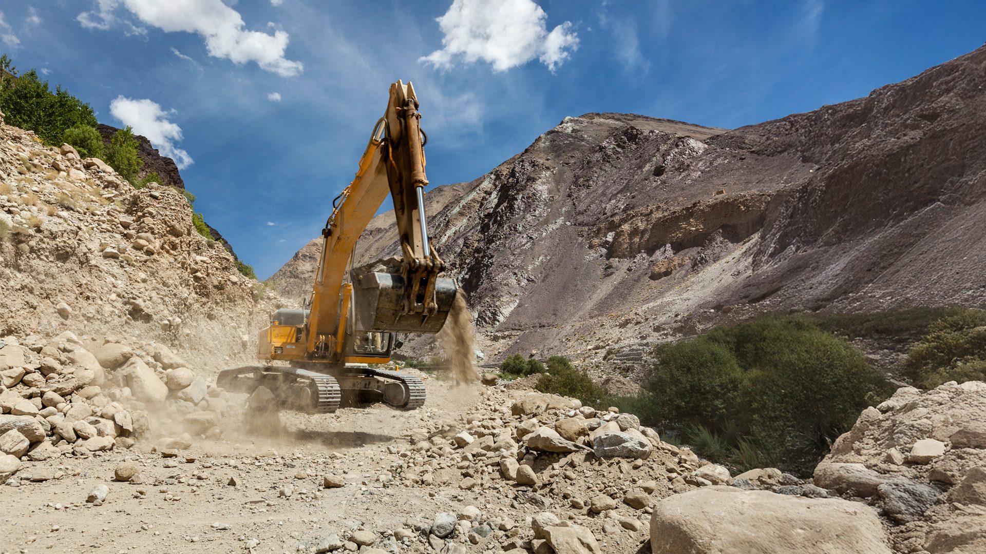 Yellow excavator moving dirt in a dusty, rocky mountain valley under a blue sky.