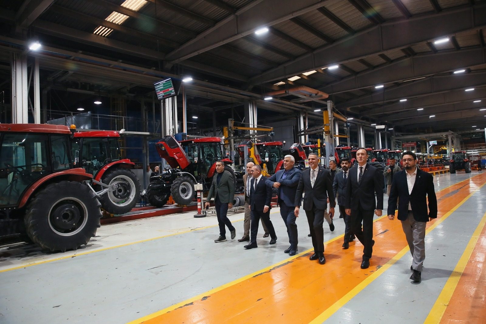 Men in suits tour a tractor factory, walking past red tractors on an assembly line.