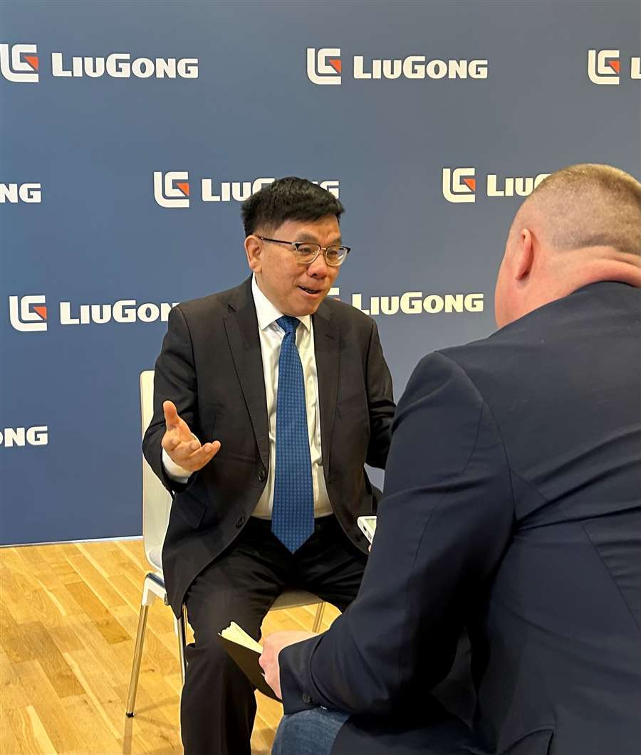 Man in suit interviewed, LiuGong branding on blue wall.