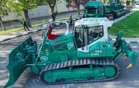 Green Liebherr bulldozer with blade and ripper on a road, green truck on a trailer behind it.
