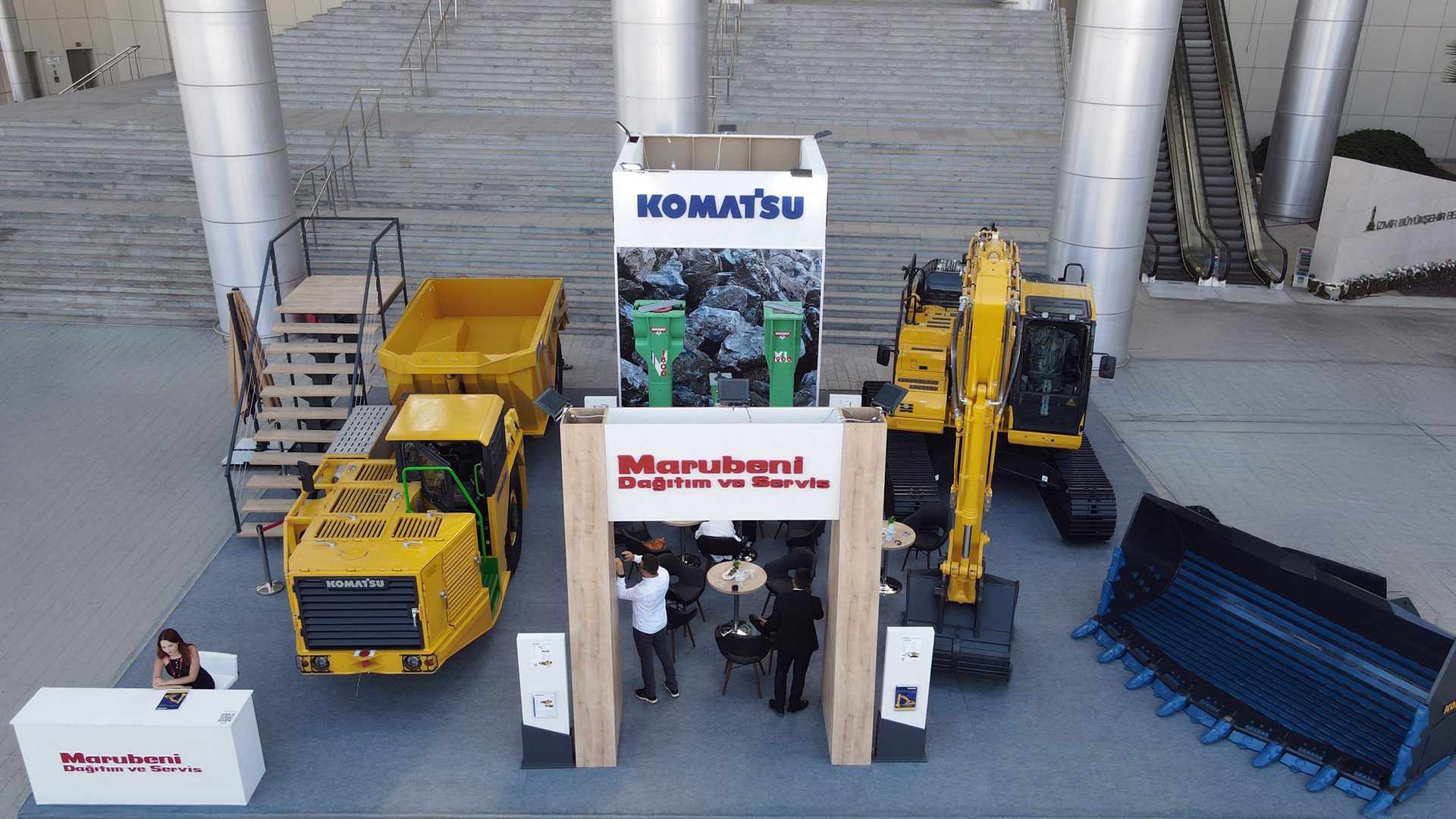 Komatsu excavator and dump truck on display with Marubeni branding at an outdoor event.