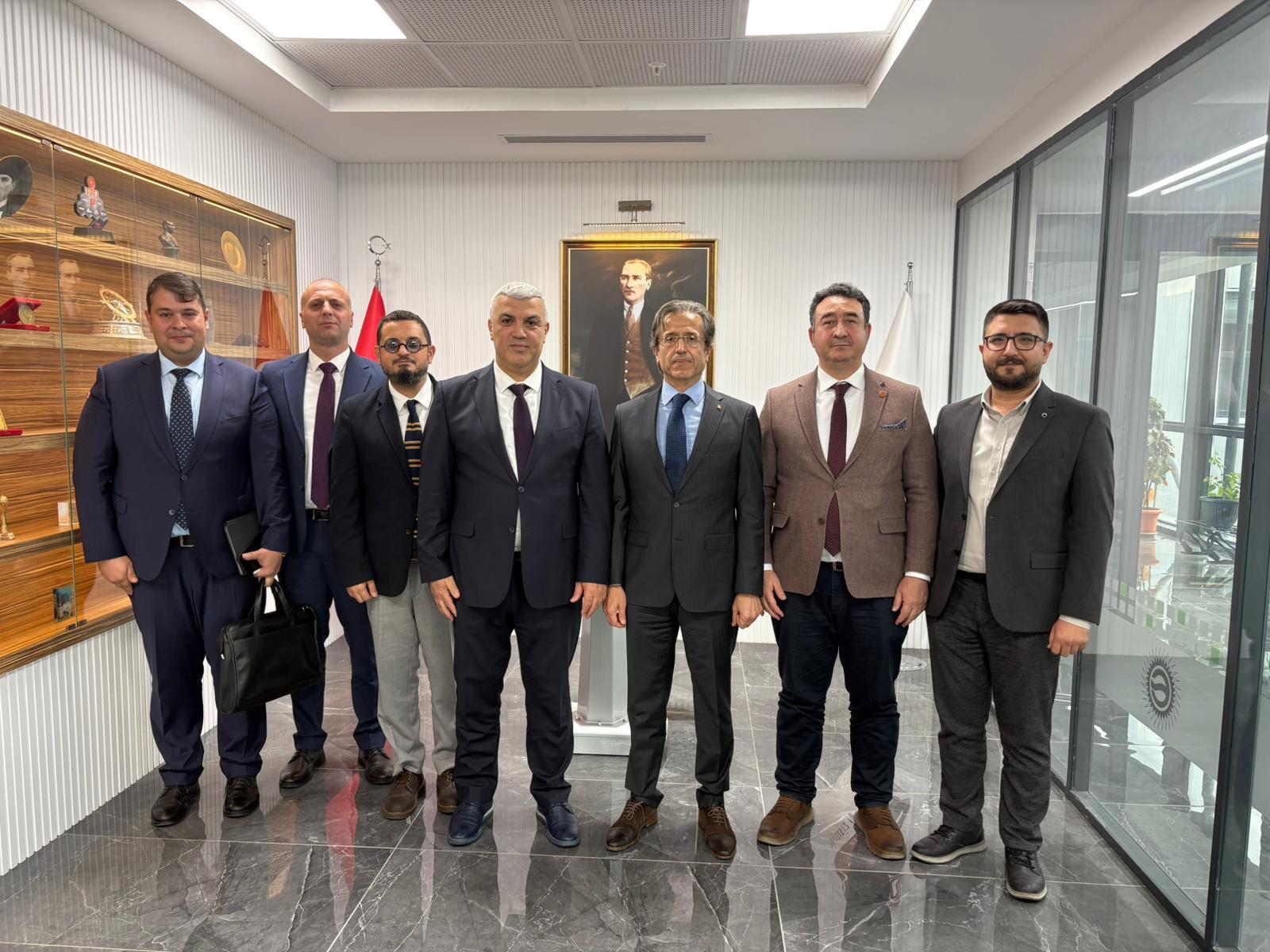 Seven men in business attire standing in a room with a portrait of a man.