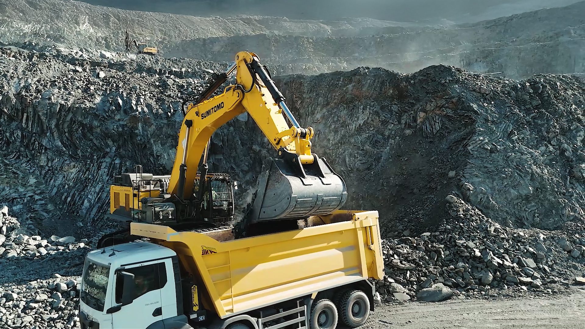 Yellow excavator loads a white truck in a rocky mine, another excavator in background.