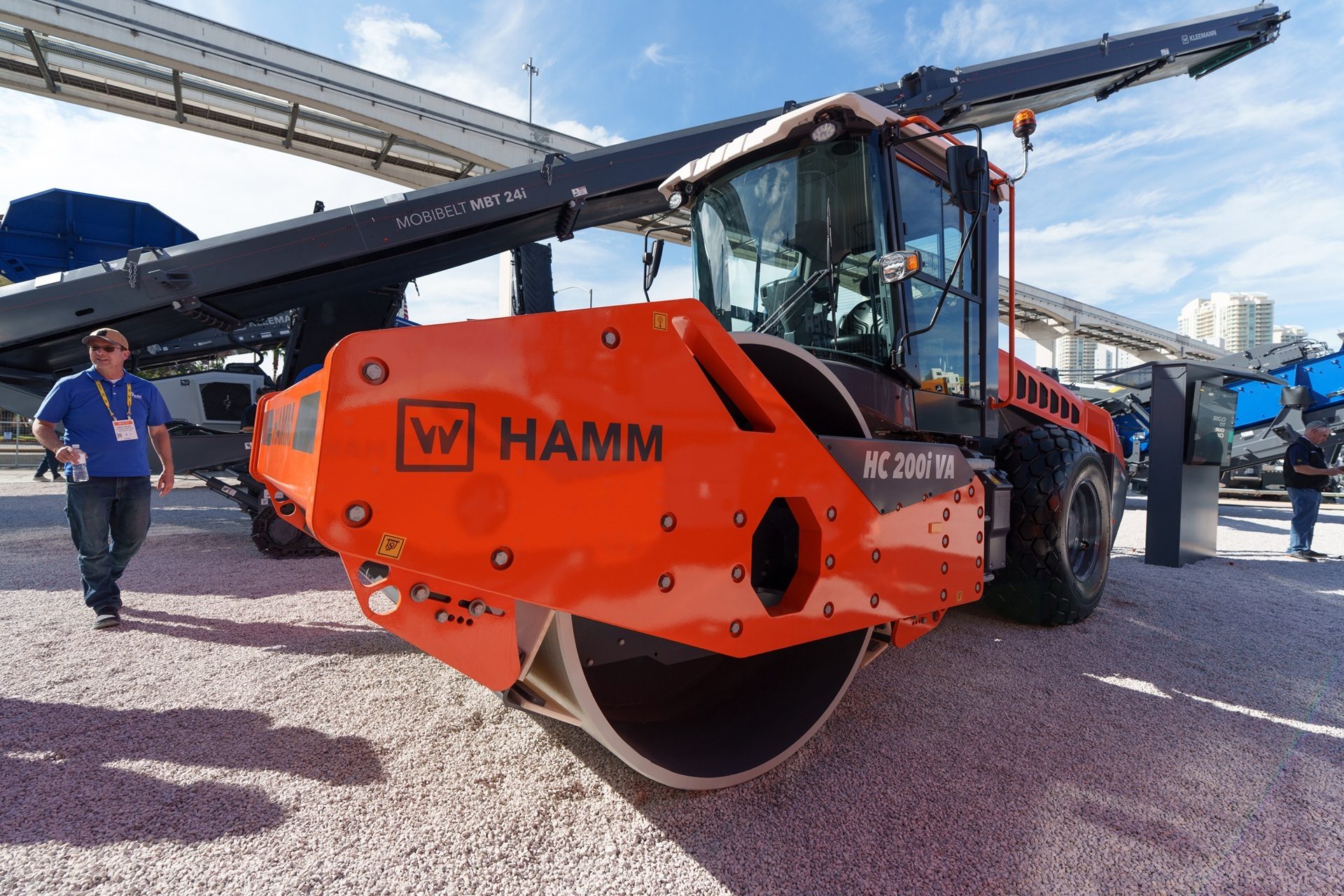 An orange HAMM HC 200i VA road roller and a MOBIBELT MBT 24i conveyor system on a gravel lot.