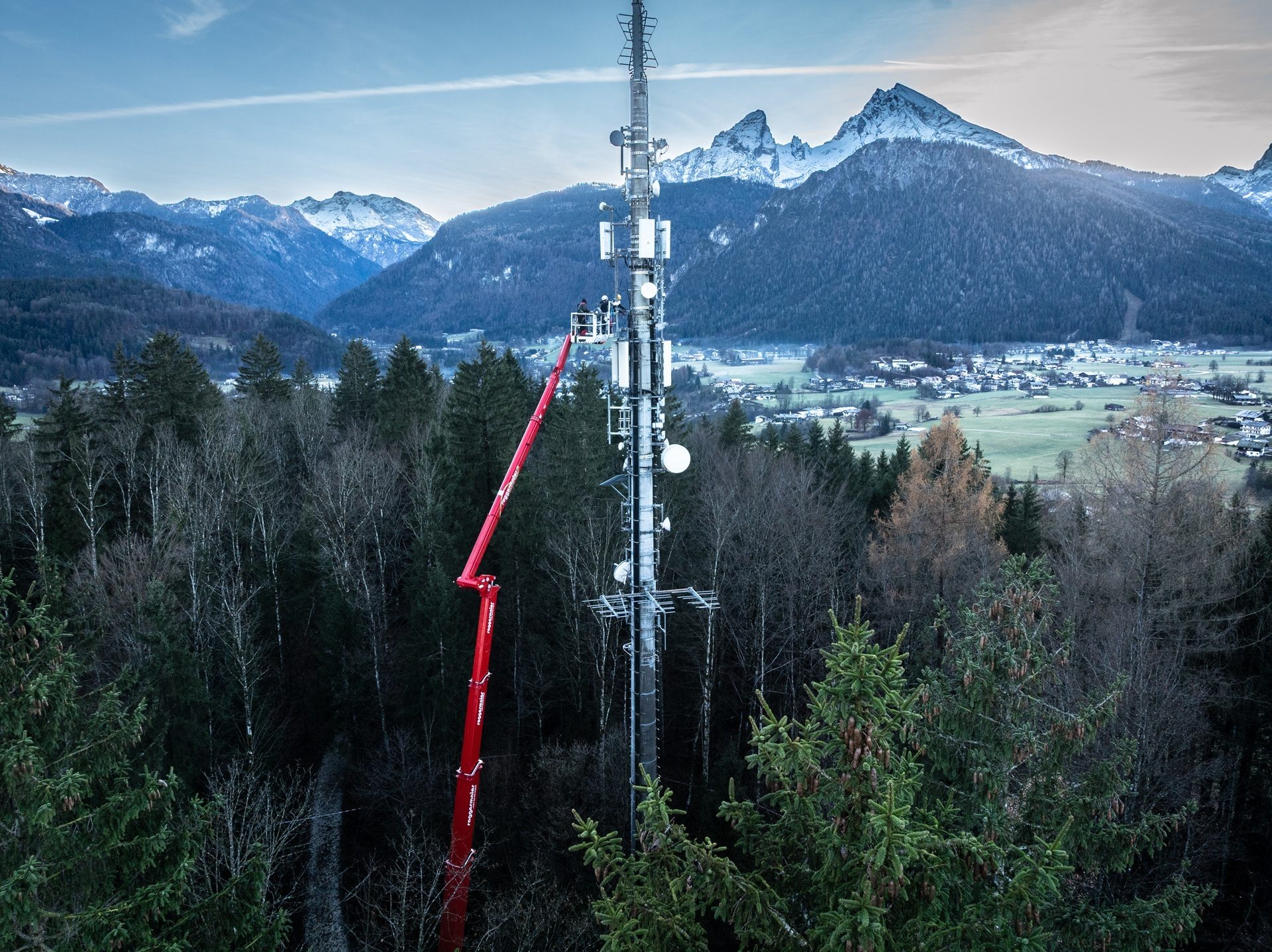 Workers on a red boom lift repair a cell tower overlooking a mountain valley and forest.