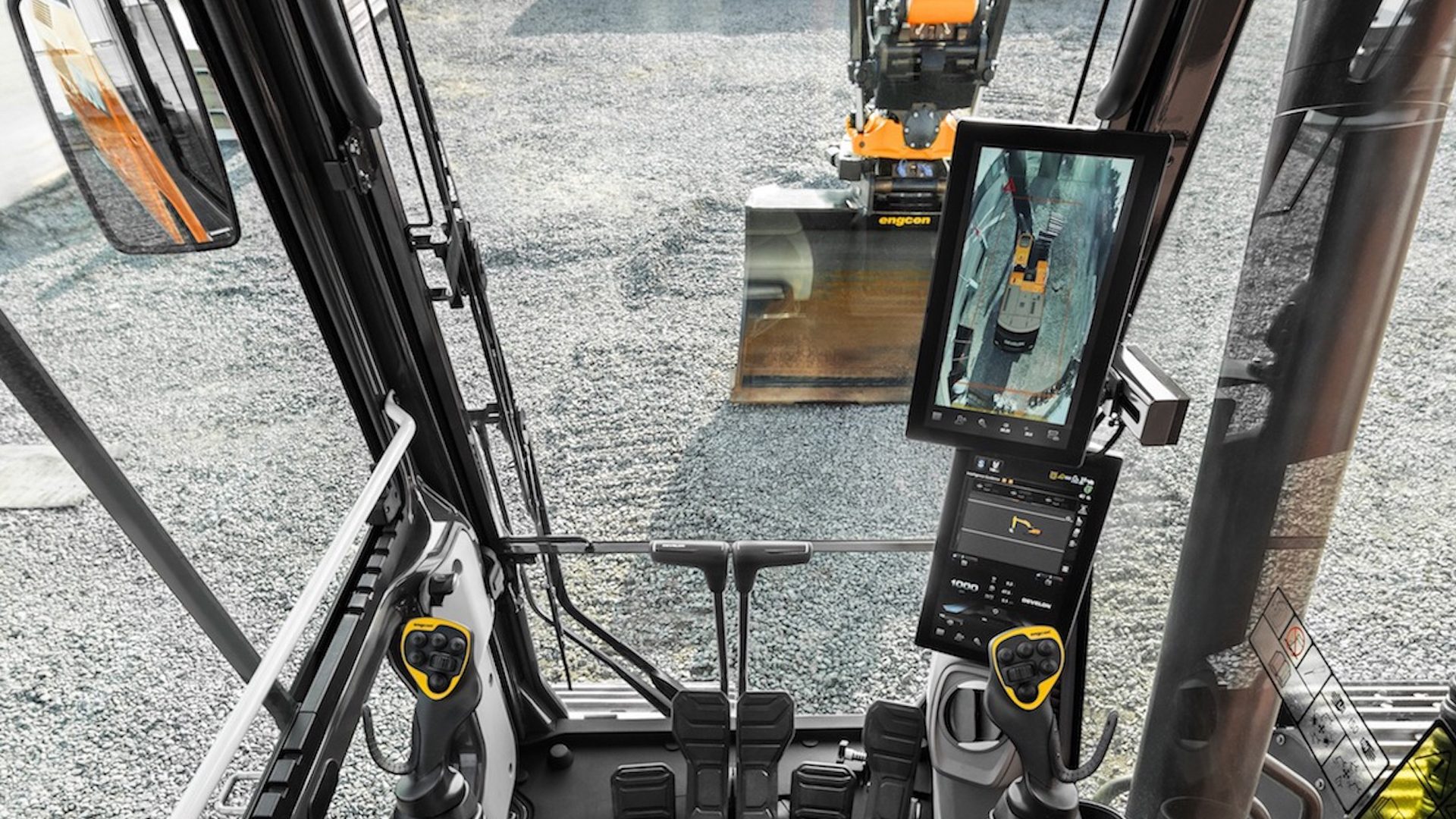 Excavator cab view: controls, two screens (camera/info), and bucket on gravel outside.