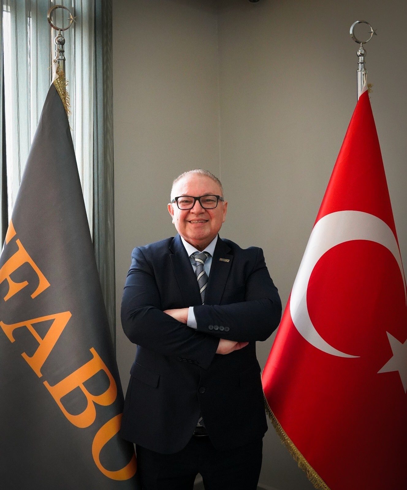 Smiling man in suit, glasses, arms crossed, between a dark flag with text and the Turkish flag.