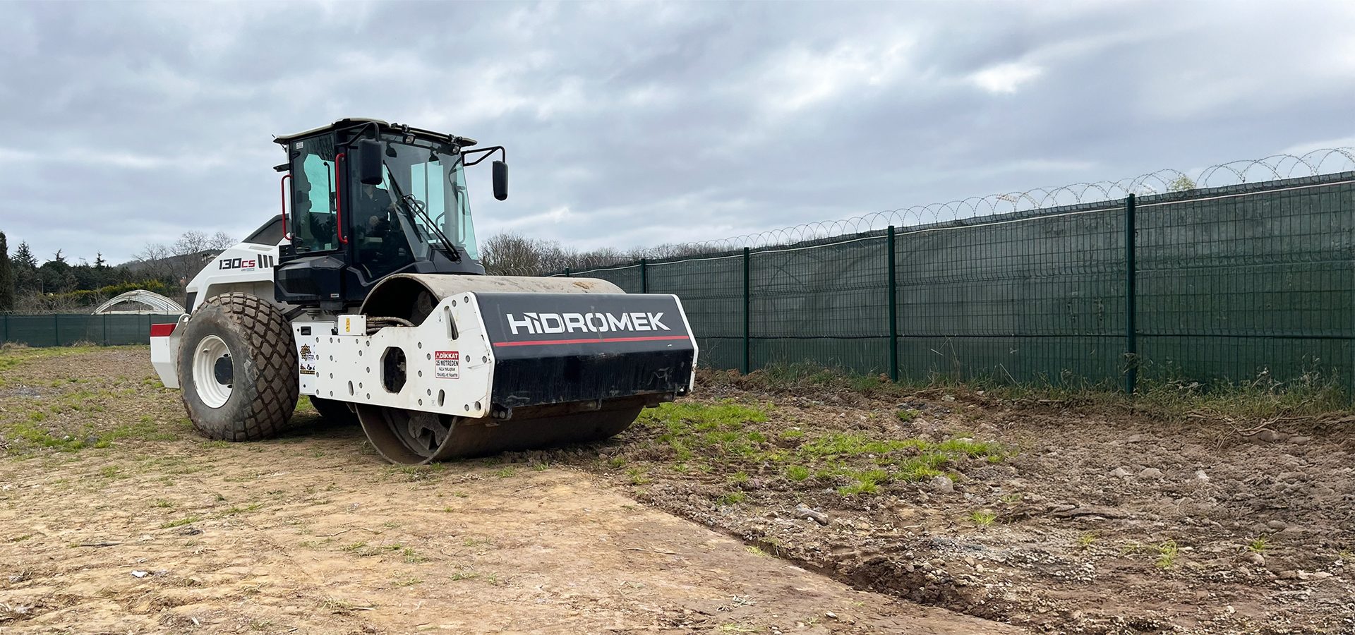 Hidromek road roller on a construction site with dirt and grass, next to a mesh fence topped with razor wire.