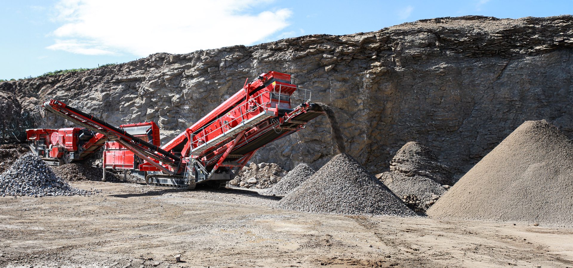 A red mobile rock crushing plant processes aggregate in a quarry, forming large piles of gravel.