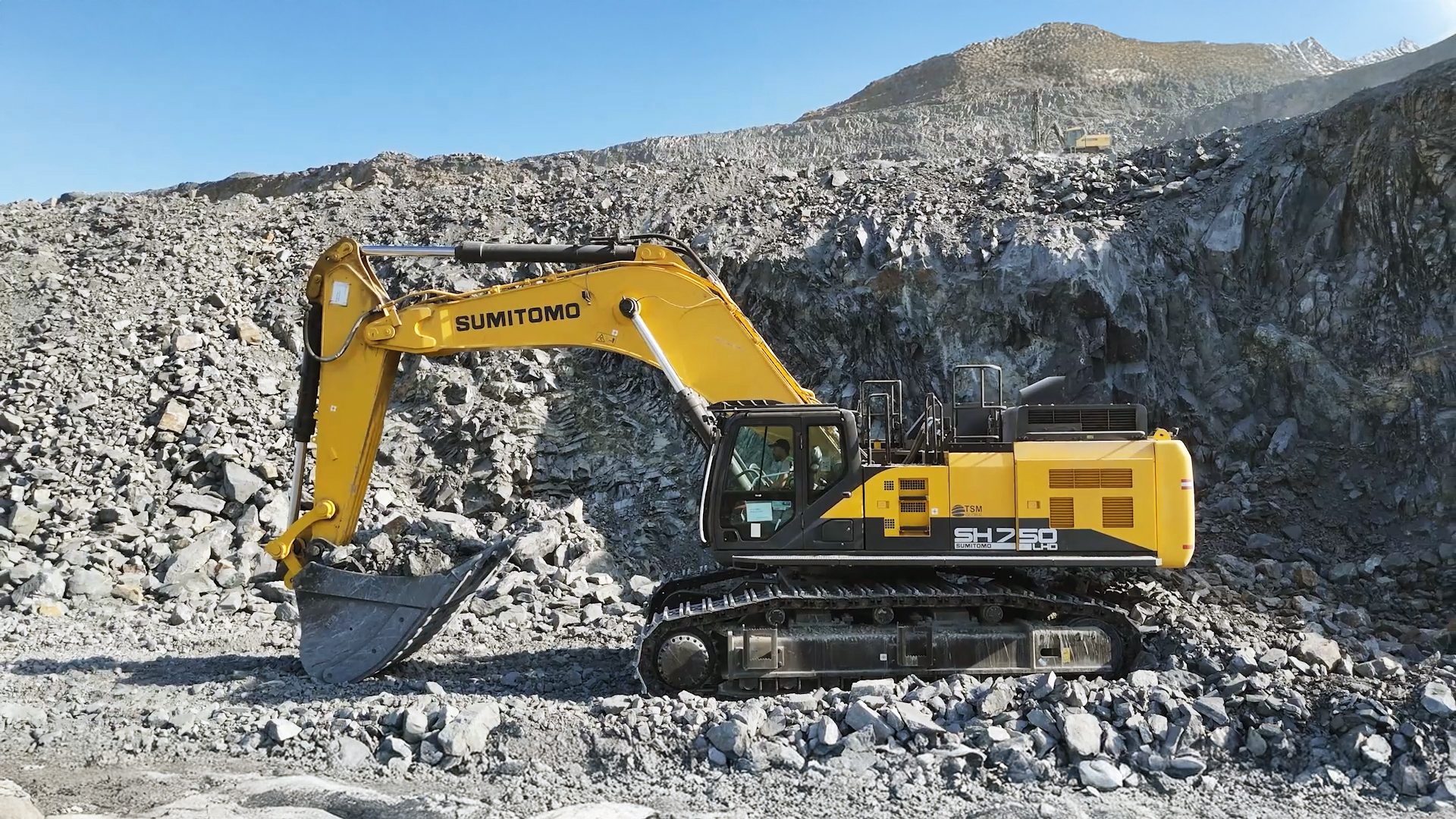 A yellow Sumitomo excavator operates in a rocky quarry under a clear sky, another excavator visible on a distant hill.