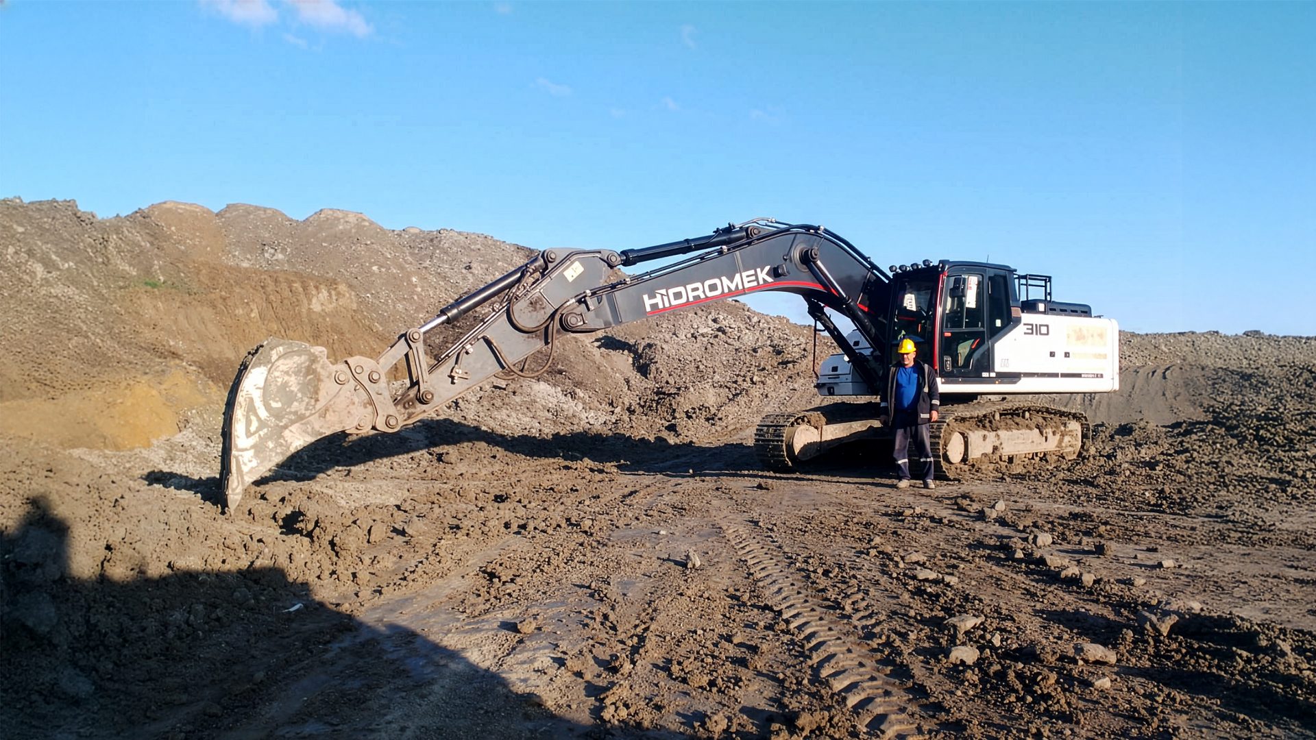 Worker by large HIDROMEK 310 excavator on a dirt construction site under a blue sky.