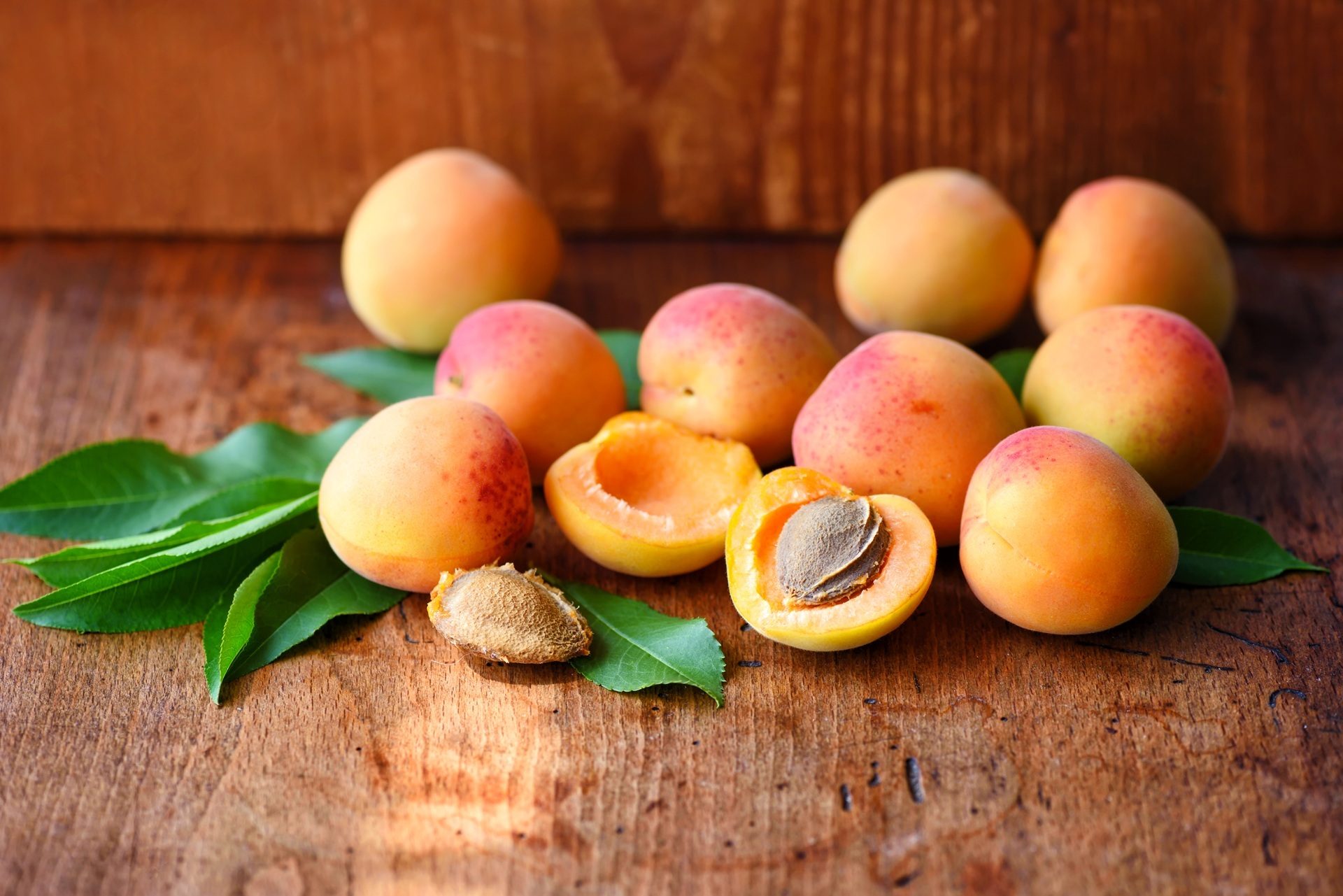Fresh apricots, some whole and some halved with pits, with green leaves on a wooden surface.