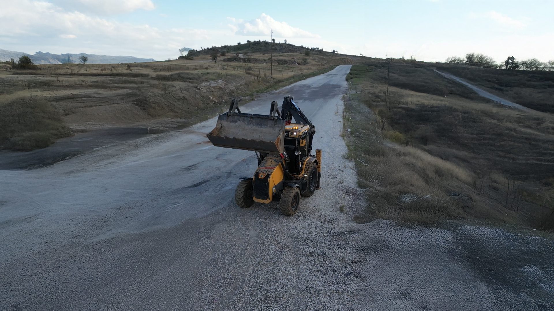 Backhoe loader with raised bucket on gravel road in hilly landscape under cloudy sky.