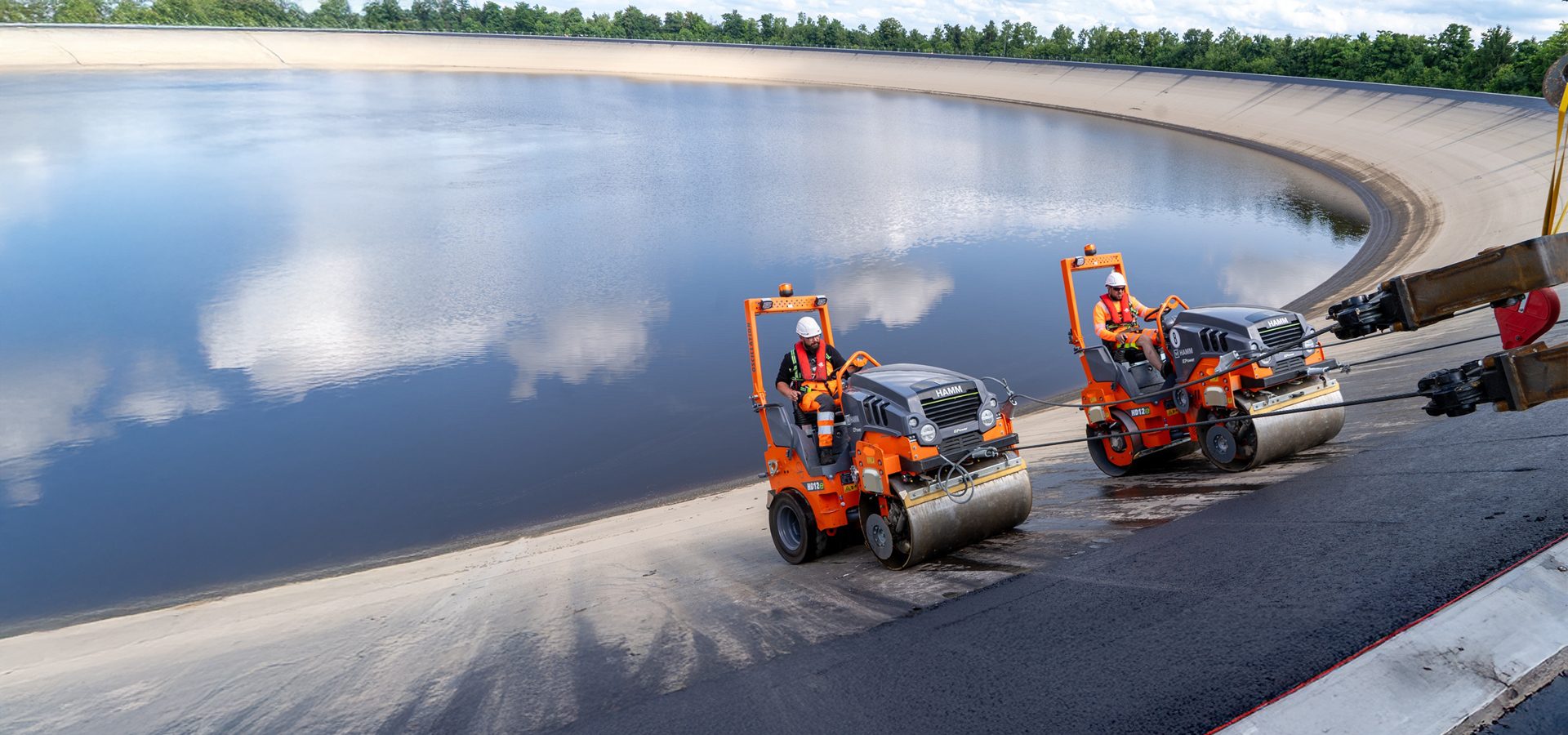 Two orange rollers pave a sloped reservoir wall, with water reflecting clouds.