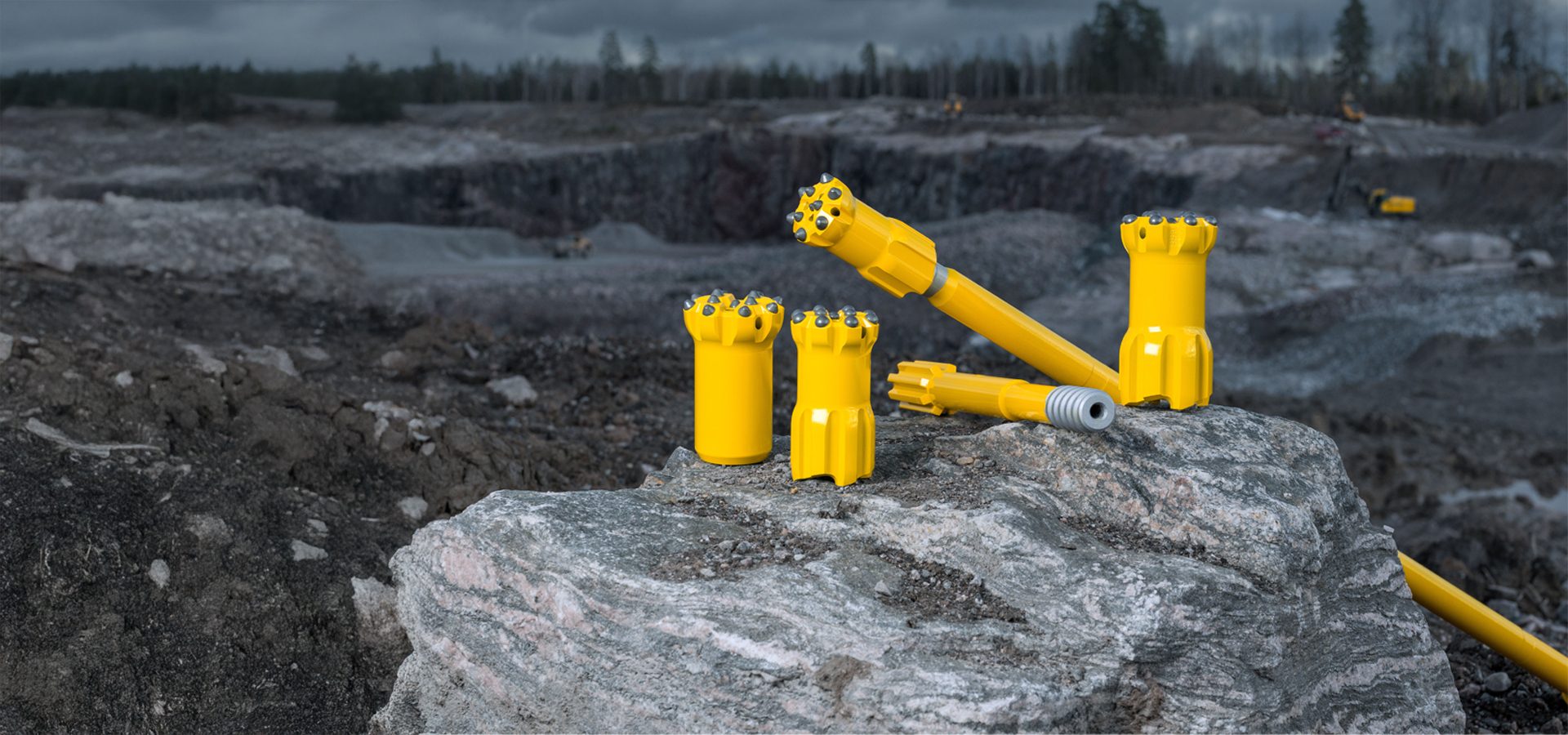 Yellow mining drill bits and rods displayed on a rock at a quarry site under a cloudy sky.