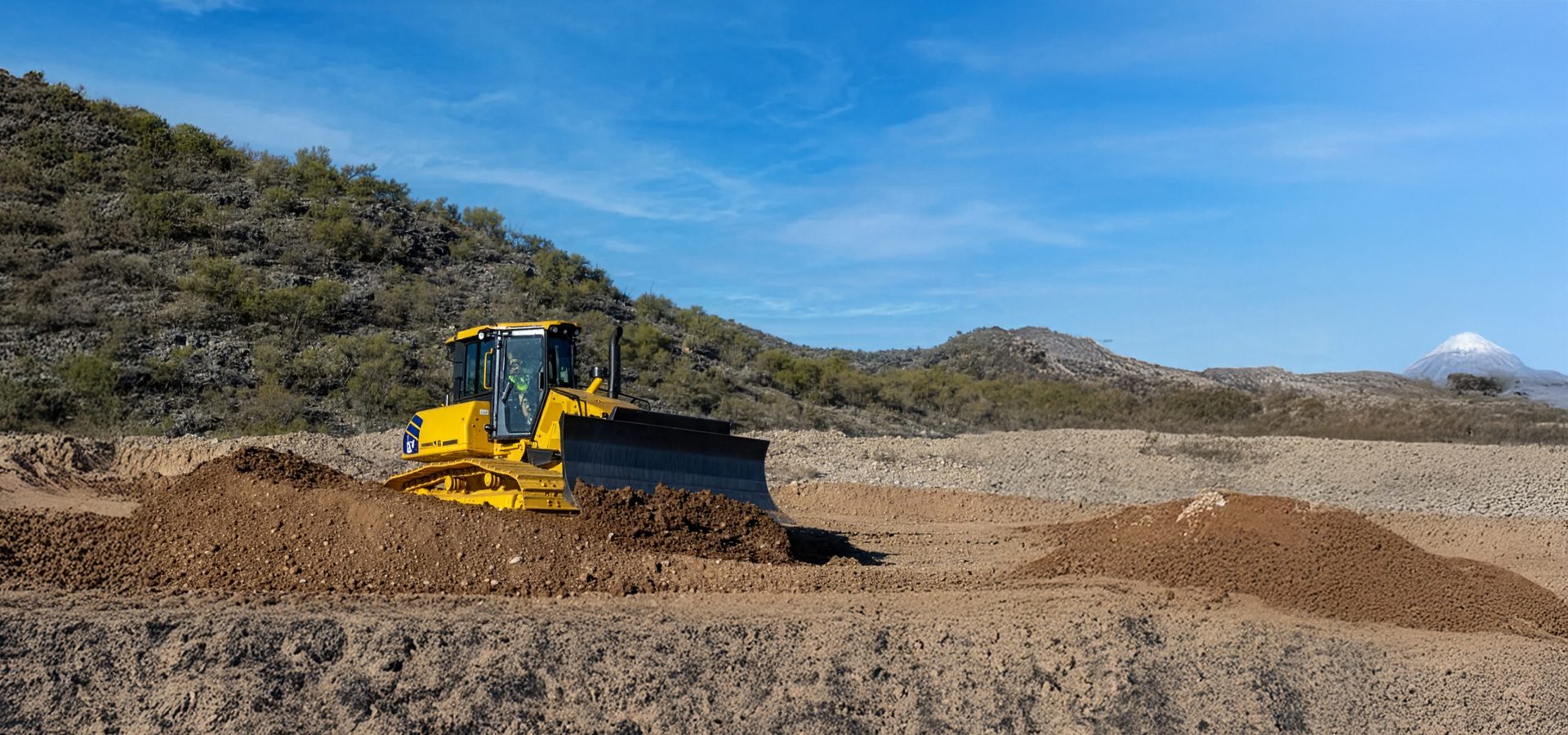 A yellow bulldozer pushes dirt on a construction site with hills and a snowy mountain in the background.