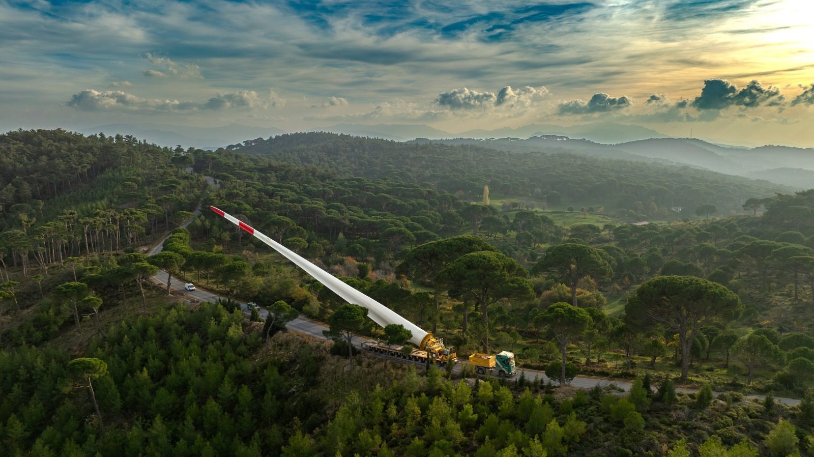 Massive wind turbine blade transported by truck through a winding road in a dense forest.