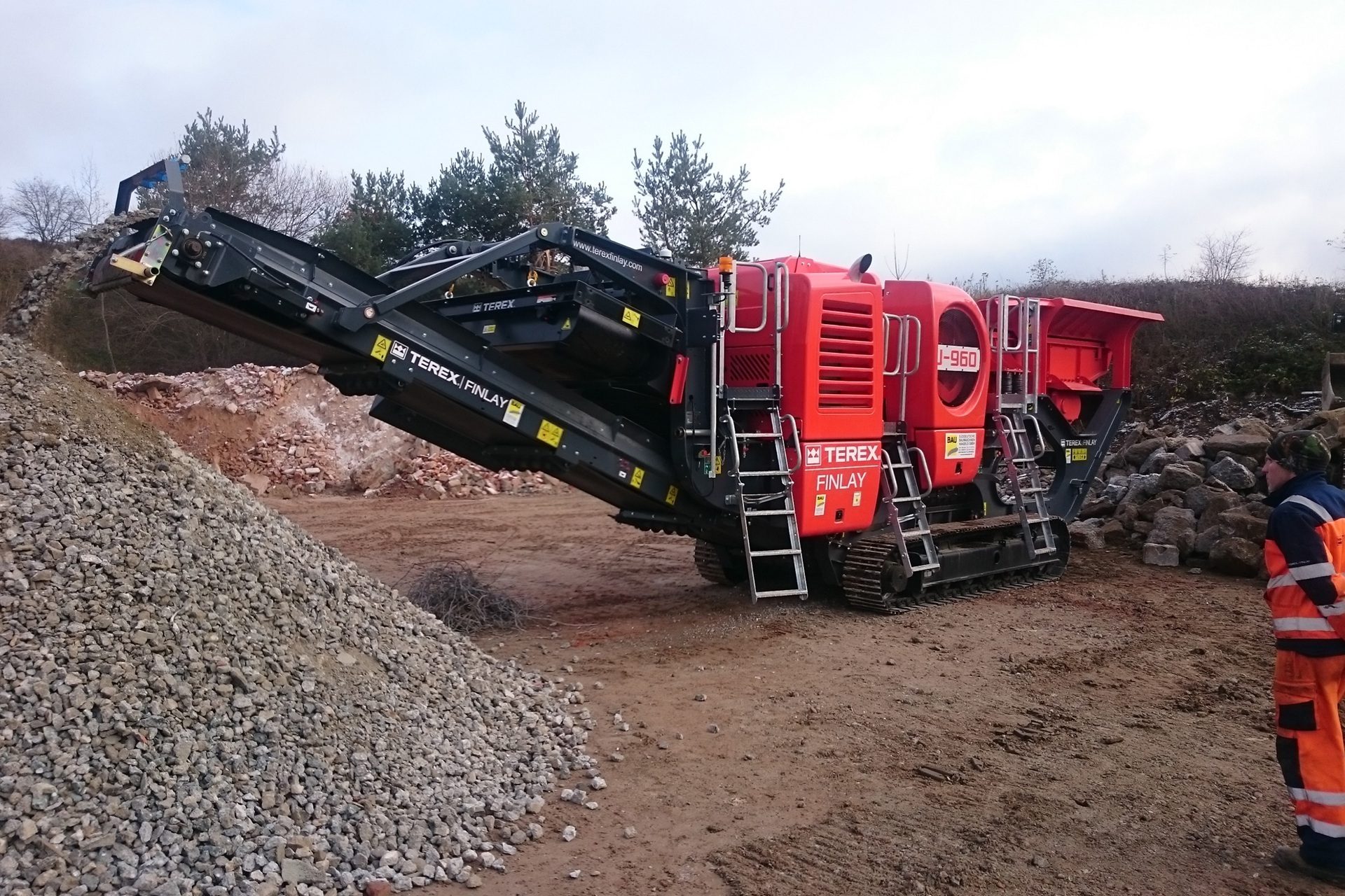 A large red and black Terex Finlay mobile crusher processes material, with a pile of aggregate and a worker.
