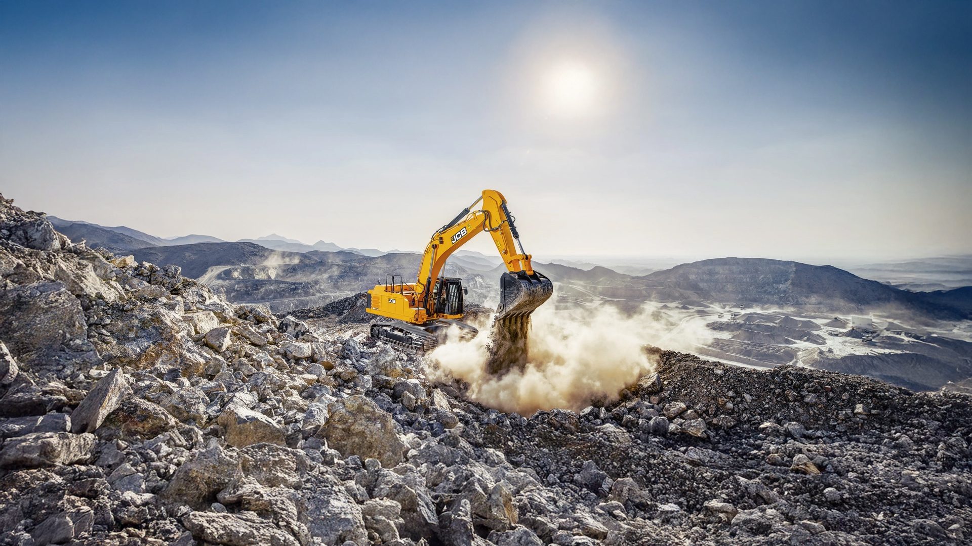 A powerful yellow JCB excavator digs into a rocky terrain, generating a cloud of dust under a clear, bright sky.