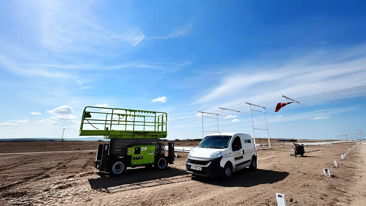 Green scissor lift, white van on dirt ground, metal frames, red flag, blue sky.