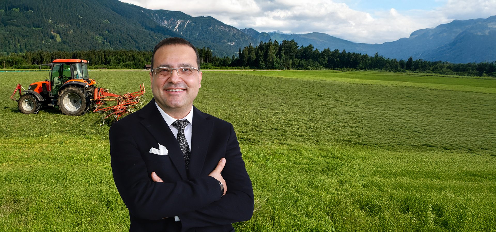 Smiling man in suit in a green farm field with a tractor and mountains.