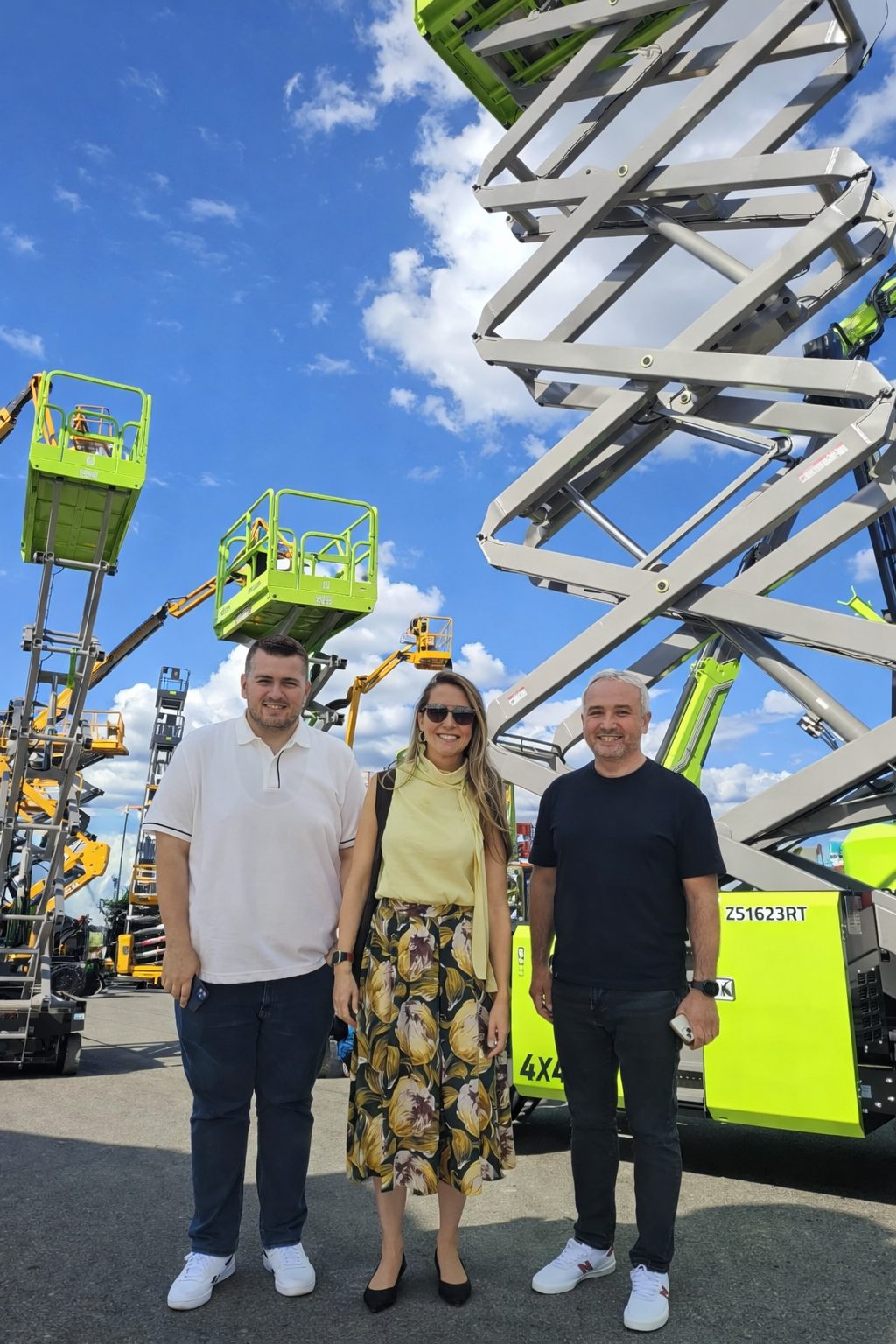 Three smiling people stand outdoors in front of multiple green and yellow scissor lifts under a blue sky.