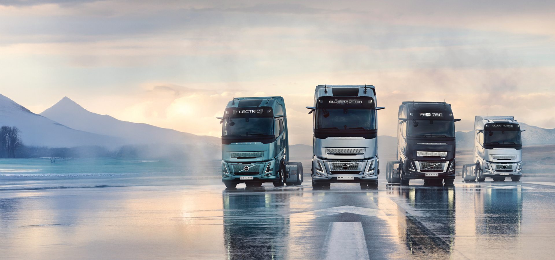 Four Volvo trucks (light blue, silver, black, white) on a wet road with mountains and sky.