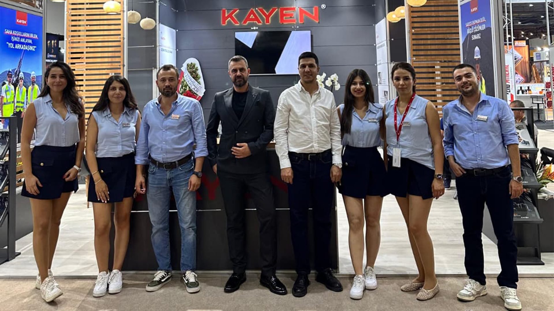 Eight people, four men and four women, smile in front of a KAYEN exhibition booth.