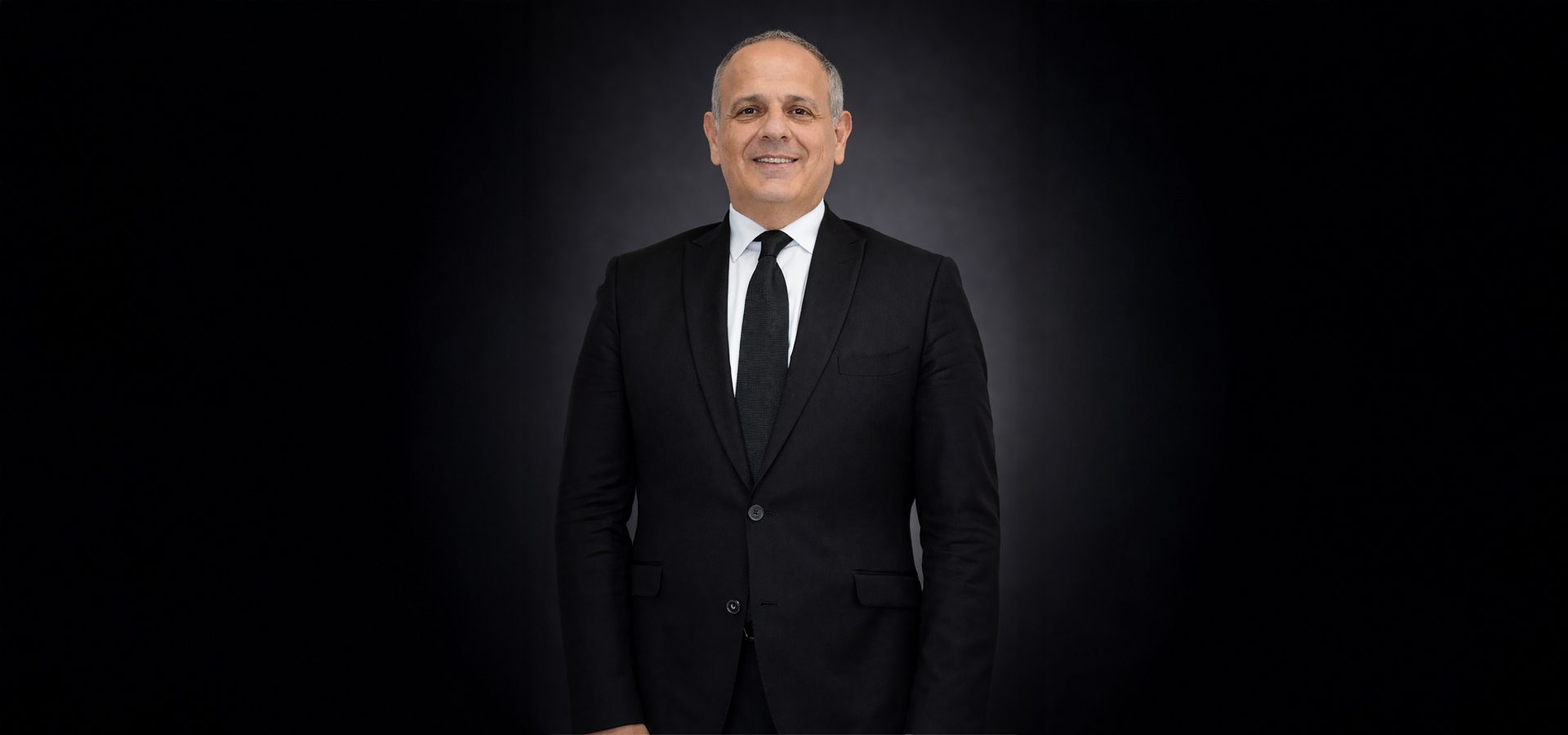 A smiling man in a black suit and tie, standing against a dark background, looking directly at the camera.