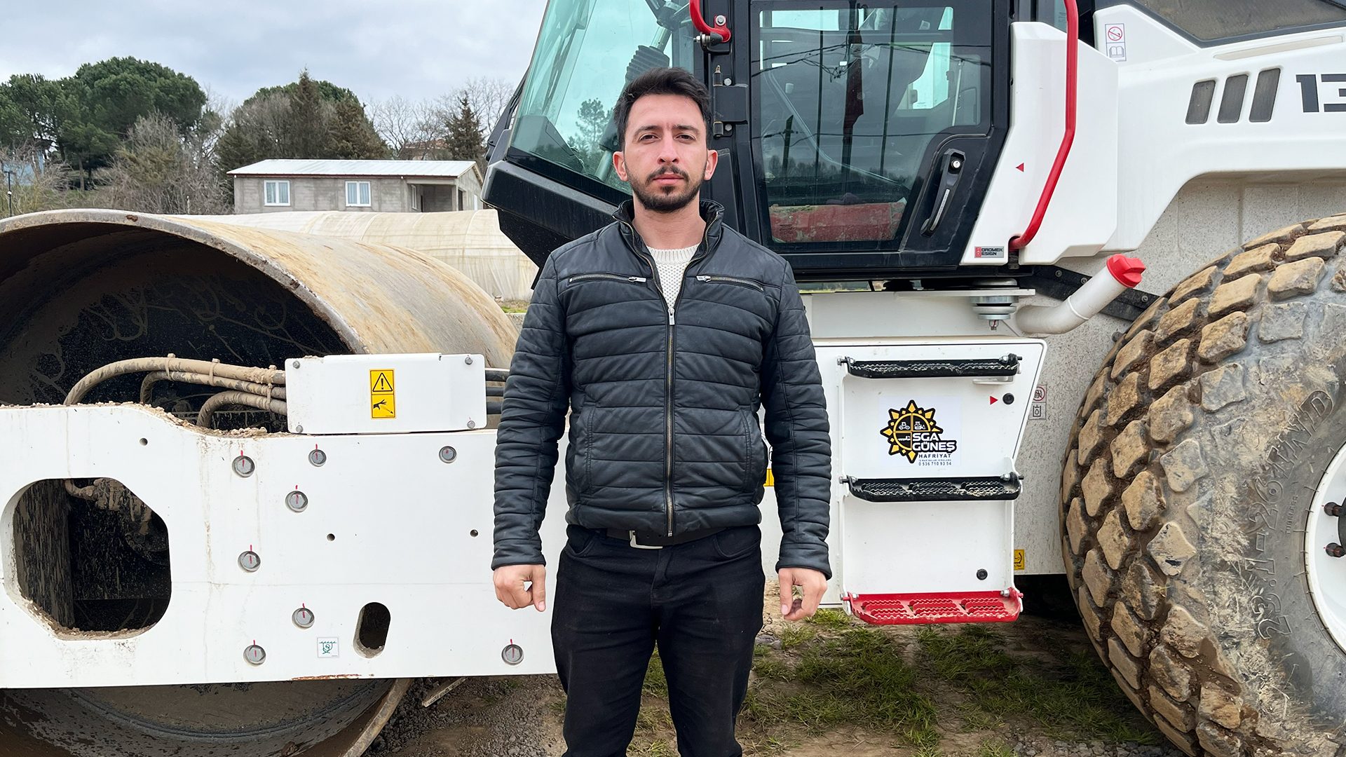 A man in a black leather jacket stands between a large road roller's drum and cabin.