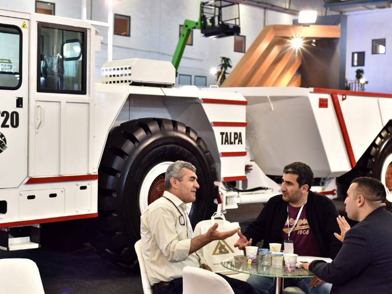 Three men discuss at a table with a large white "TALPA" mining truck in the background at an exhibition.