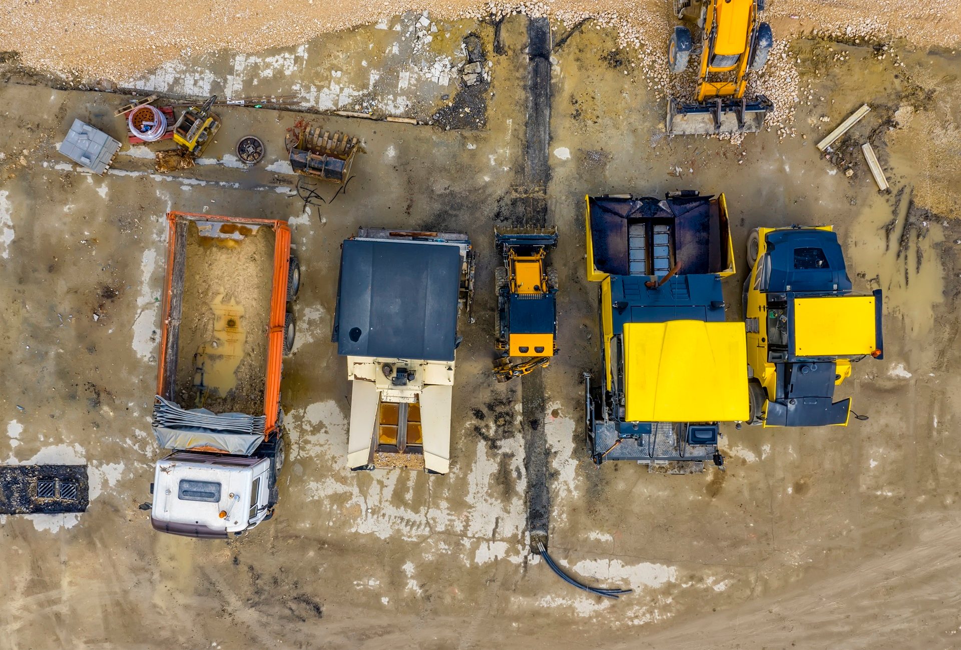 Aerial view of a muddy construction site with heavy machinery: trucks, excavators, and pavers.