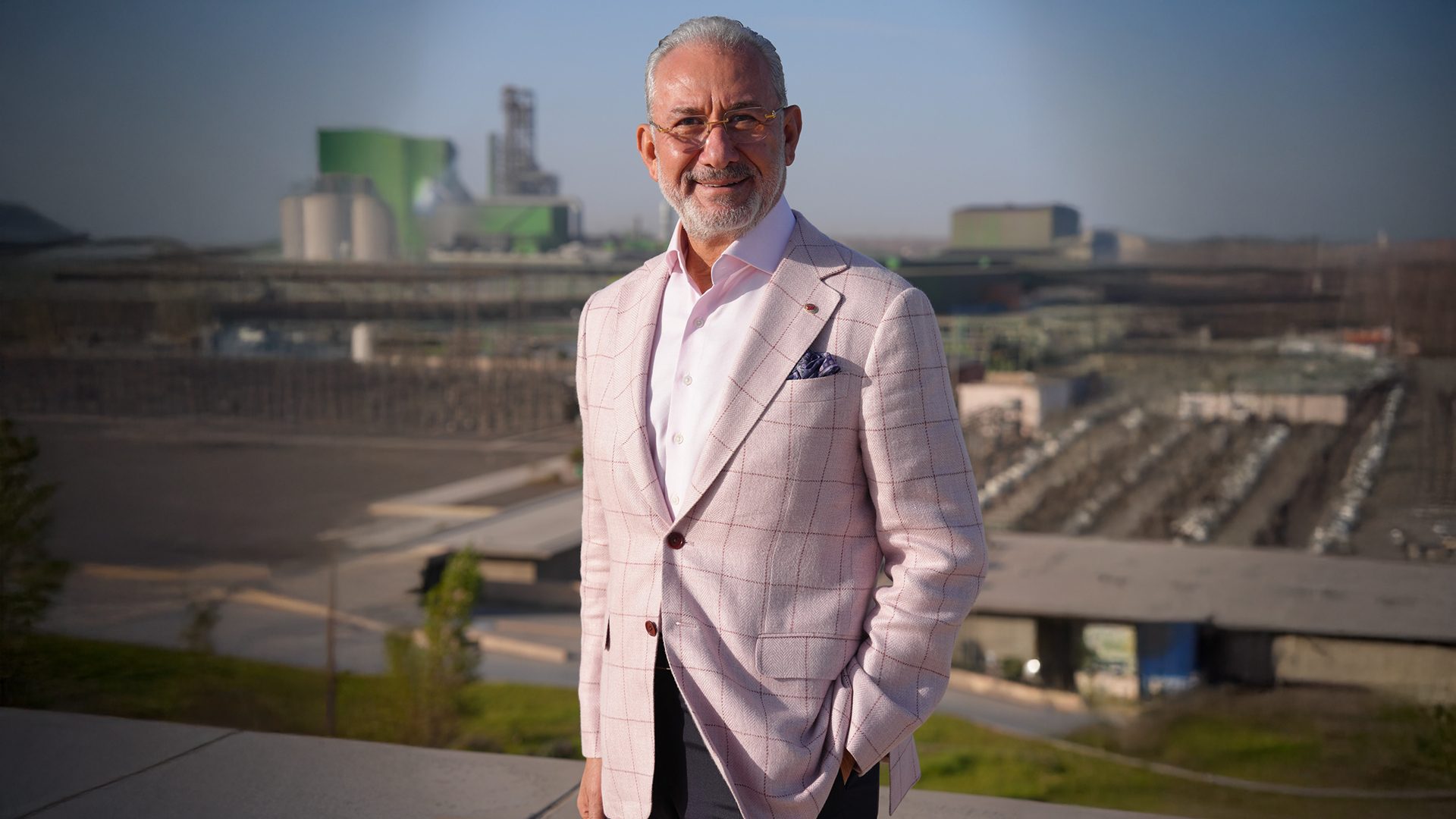 Smiling man in pink plaid suit and glasses, gray hair and beard, industrial background.