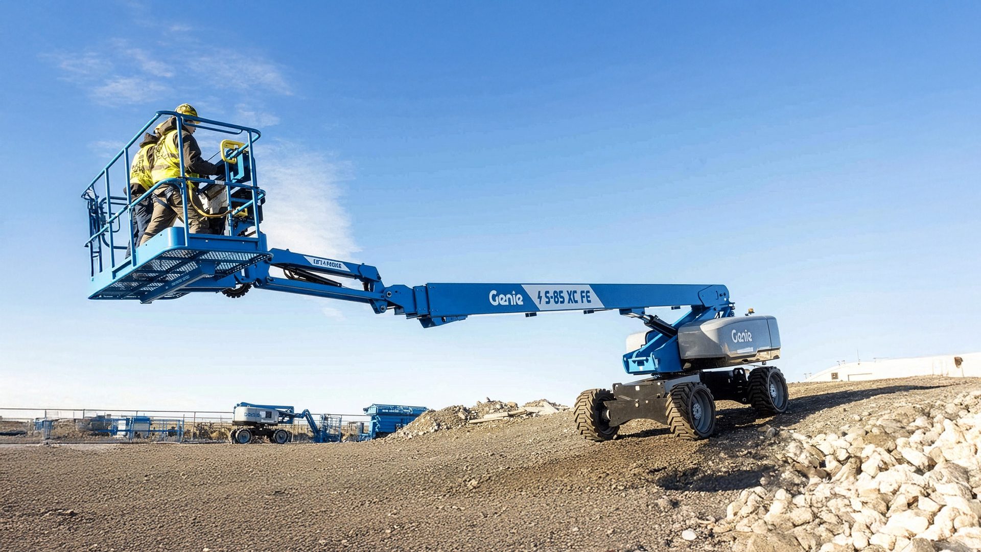Two workers in a blue Genie S-85 XC FE boom lift on a construction site under a clear sky.