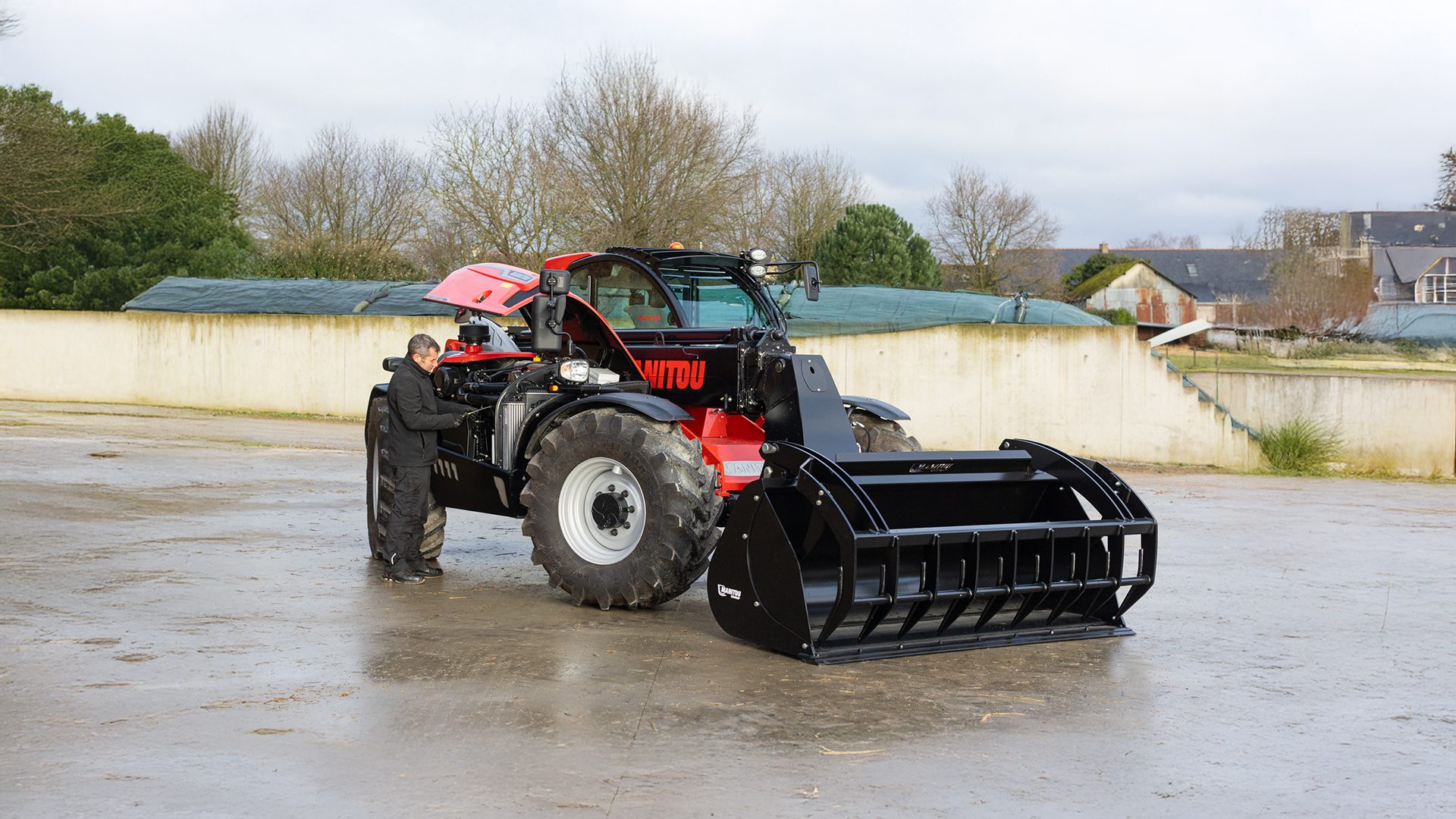Man checking a red and black Manitou telehandler with a large bucket attachment on a wet farmyard.