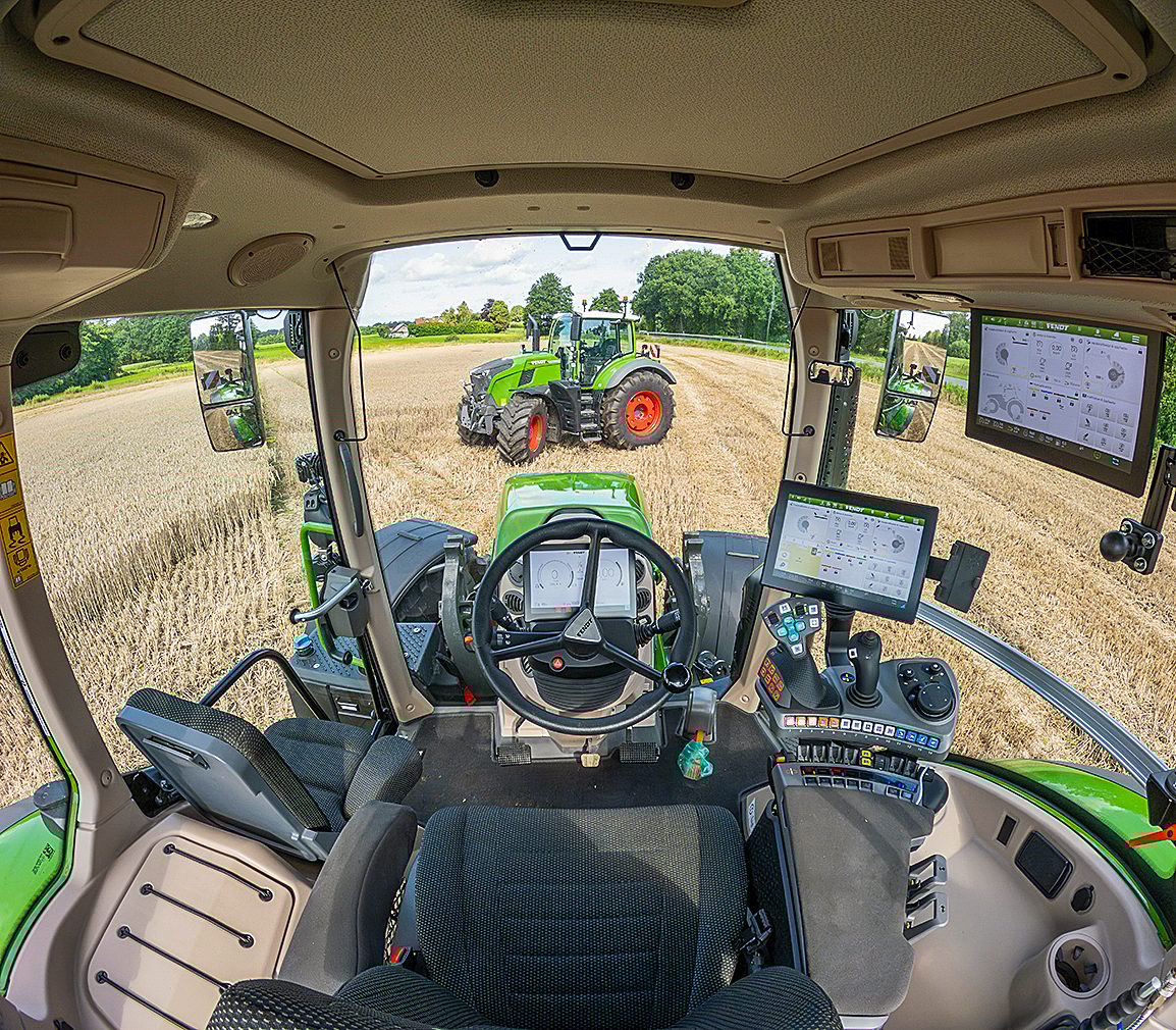 Modern tractor cab interior, controls visible, looking at another tractor in a harvested field.