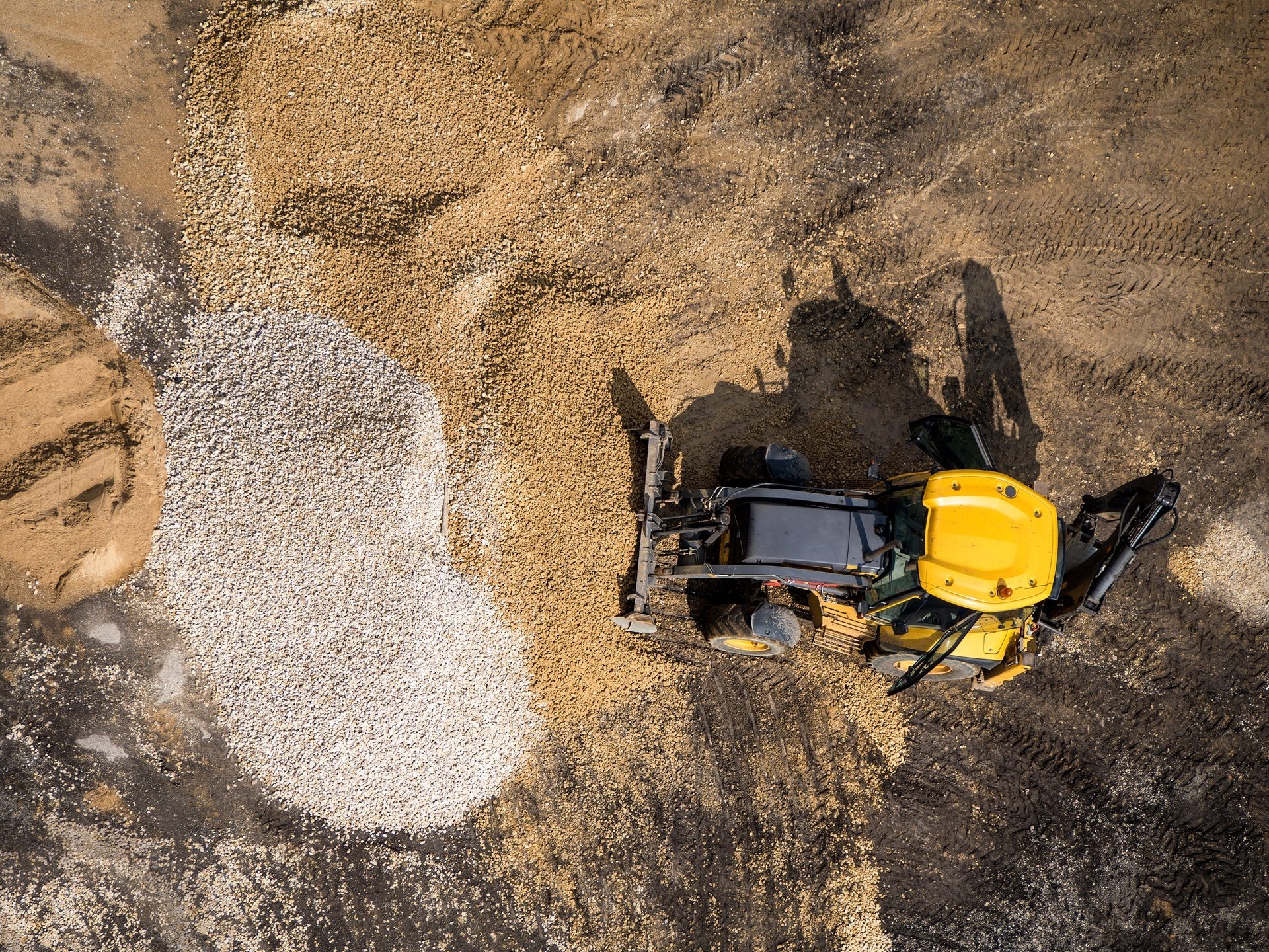 Aerial view of a yellow excavator on a construction site with piles of gravel and sand, casting shadows.